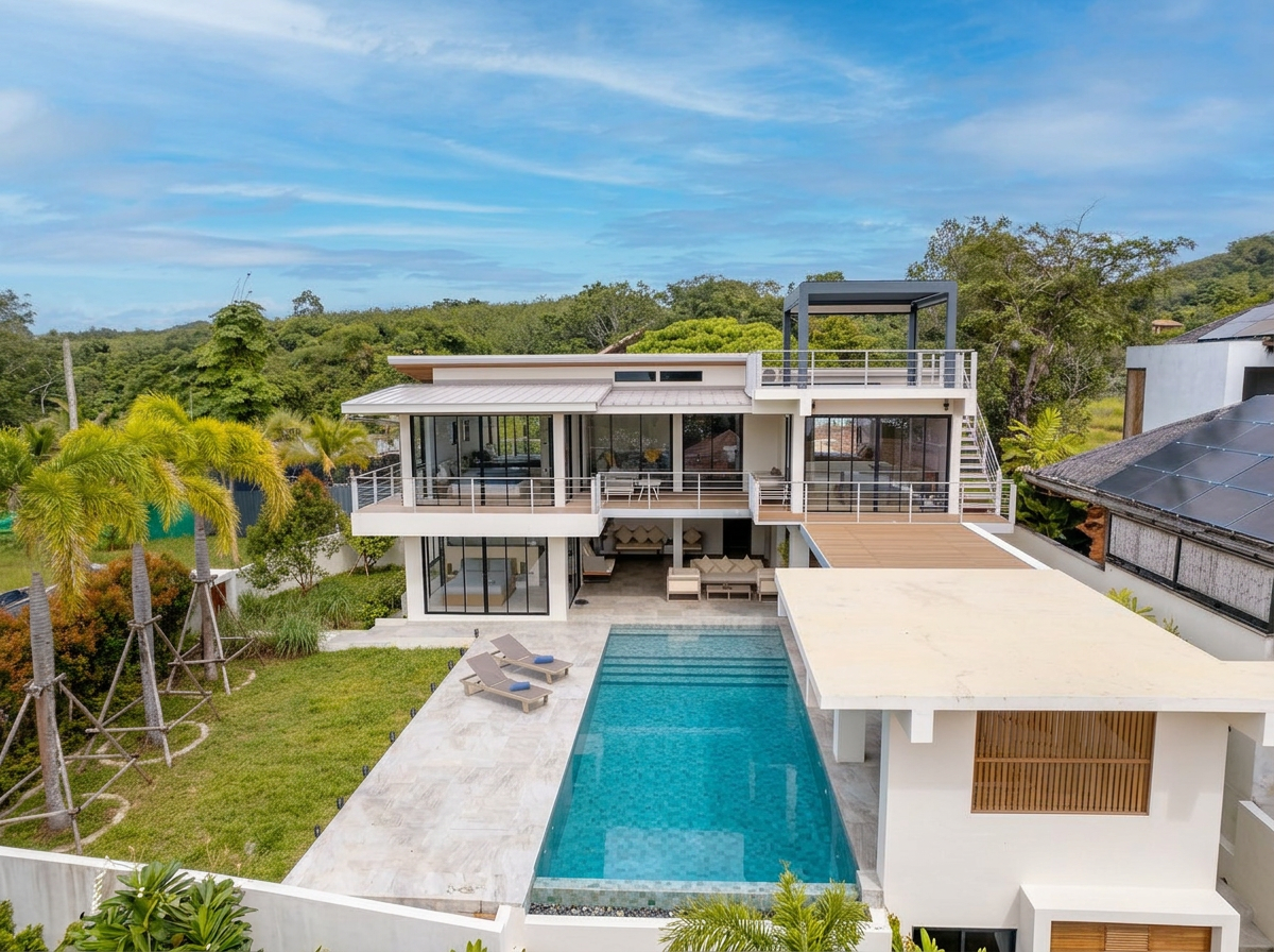 Modern house with outdoor pool, patio furniture, surrounded by lush greenery and trees, under a partly cloudy sky.