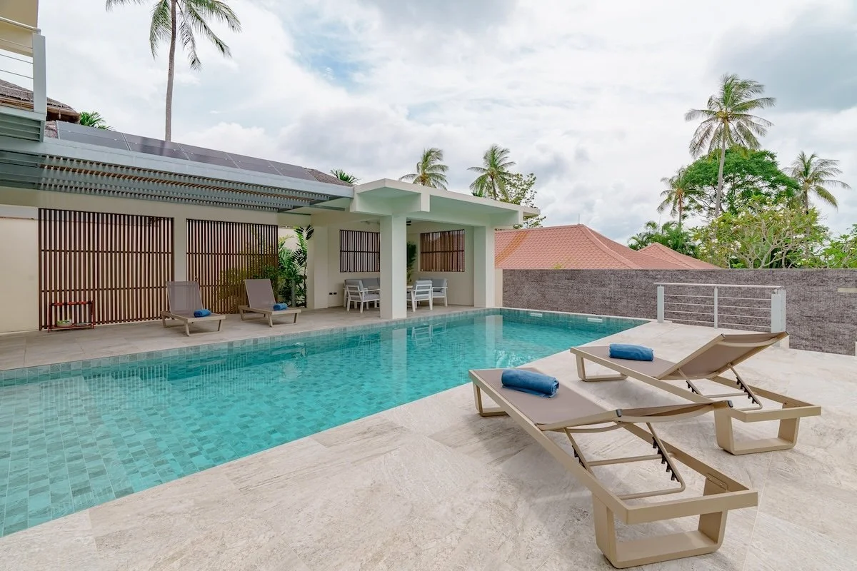 Private pool villa koh yao noi
A modern outdoor swimming pool with two beige lounge chairs and blue towels, surrounded by a patio with palm trees and a privacy wall, under a partly cloudy sky.