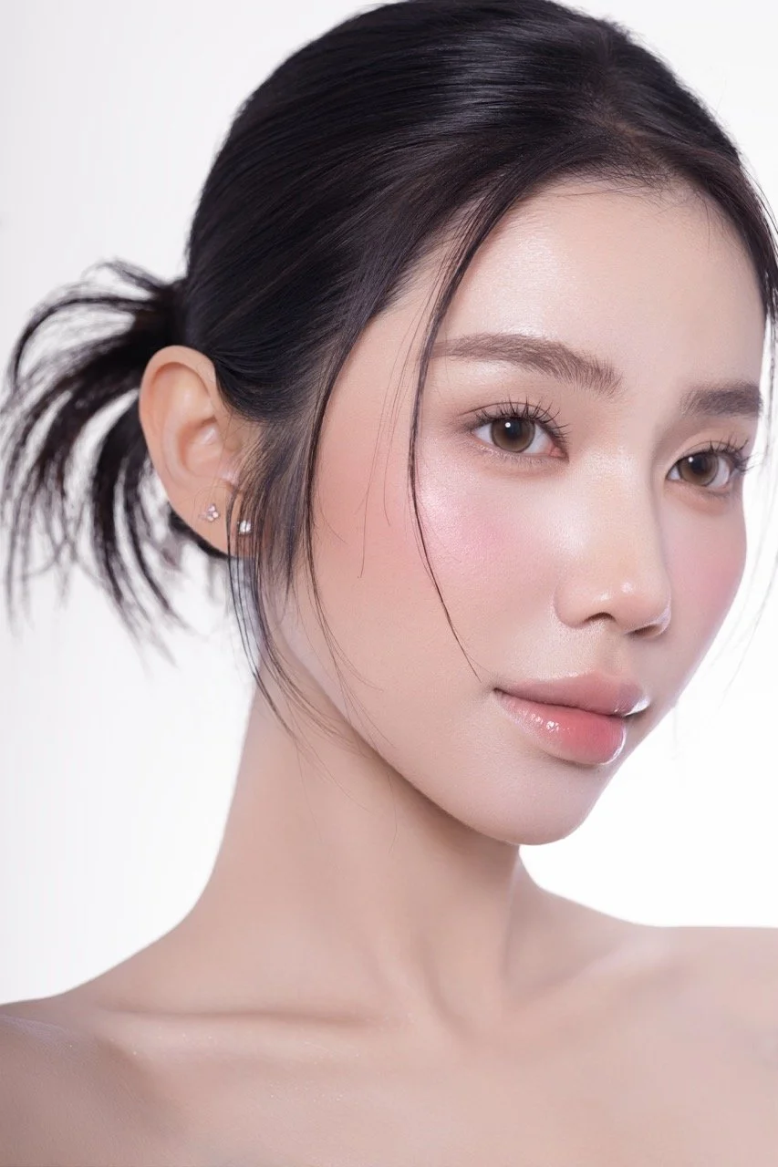 Close-up portrait of a young woman with fair skin, dark hair tied in a messy bun, light makeup, and earrings, against a plain white background.