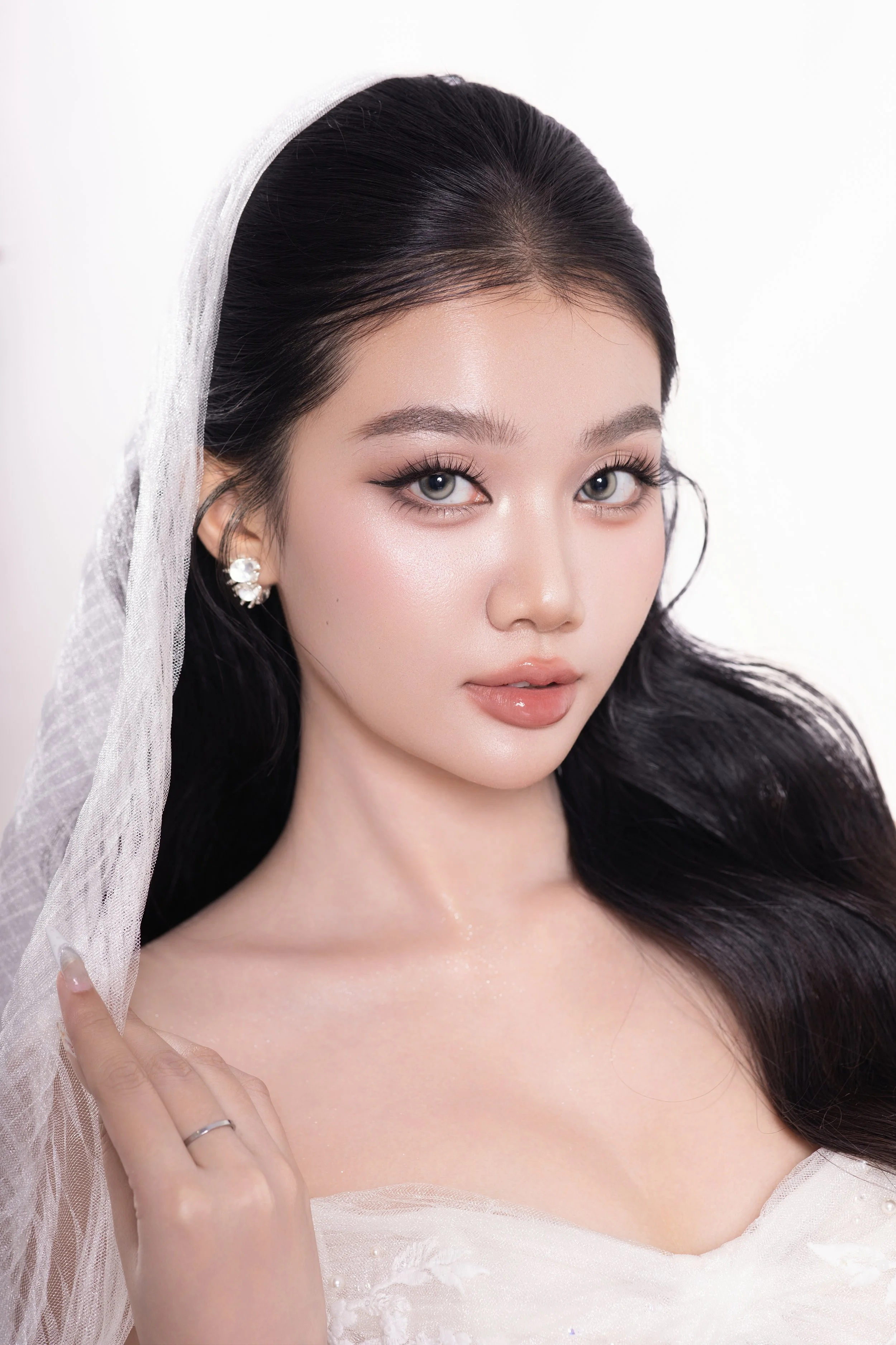 Close-up of a woman with long black hair, blue eyes, and light skin, wearing pearl earrings and a veil, suggesting a bridal portrait.
