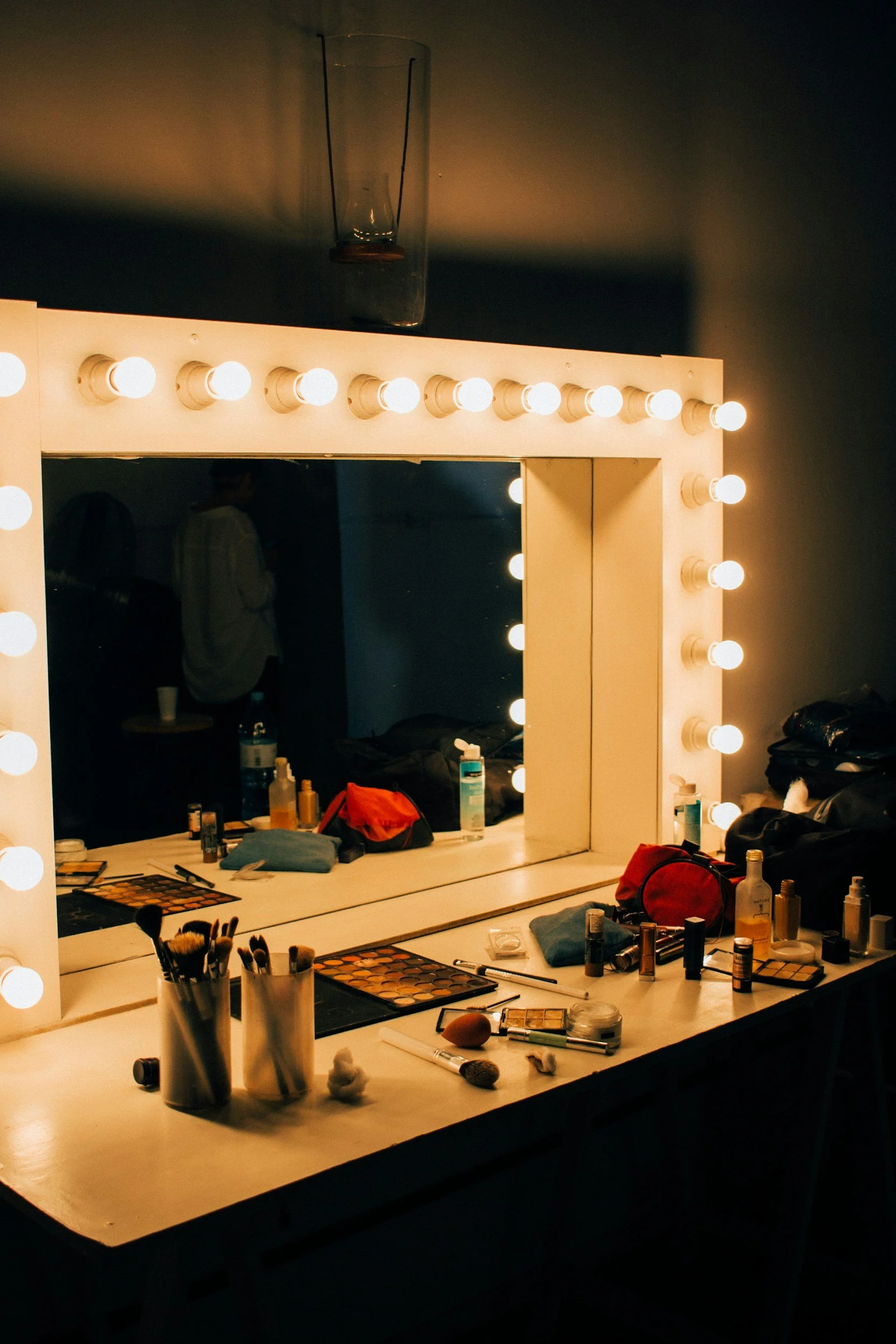 Makeup vanity mirror with bright lights, cluttered with makeup products, brushes, and personal items, reflected in a large mirror.