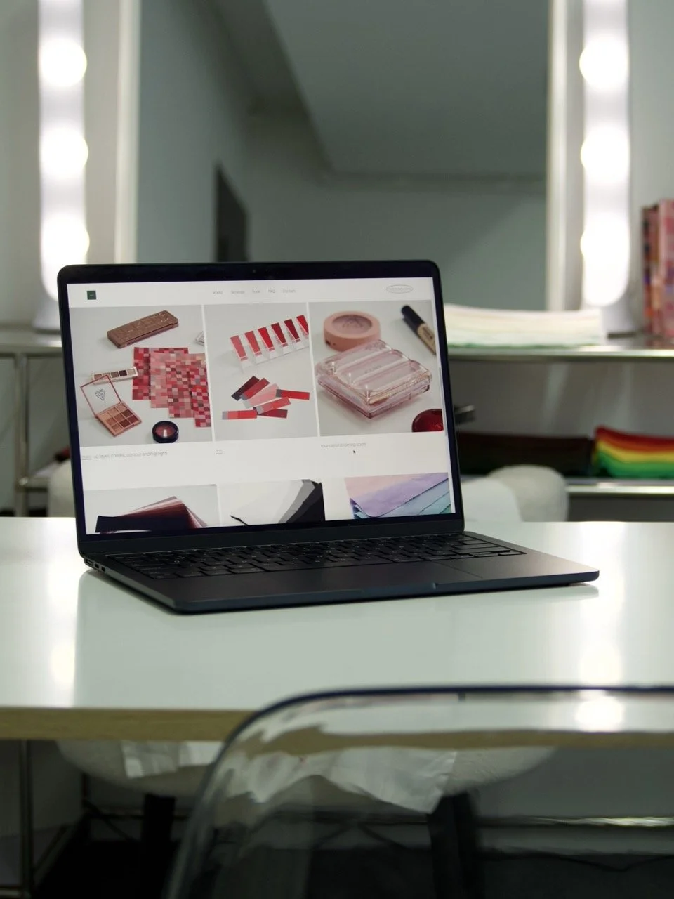 A laptop on a white table showing a webpage with various makeup and cosmetic products, including lipstick, blush, and makeup palettes. The room has a mirror with lights on either side and some shelves with papers and colorful items in the background.