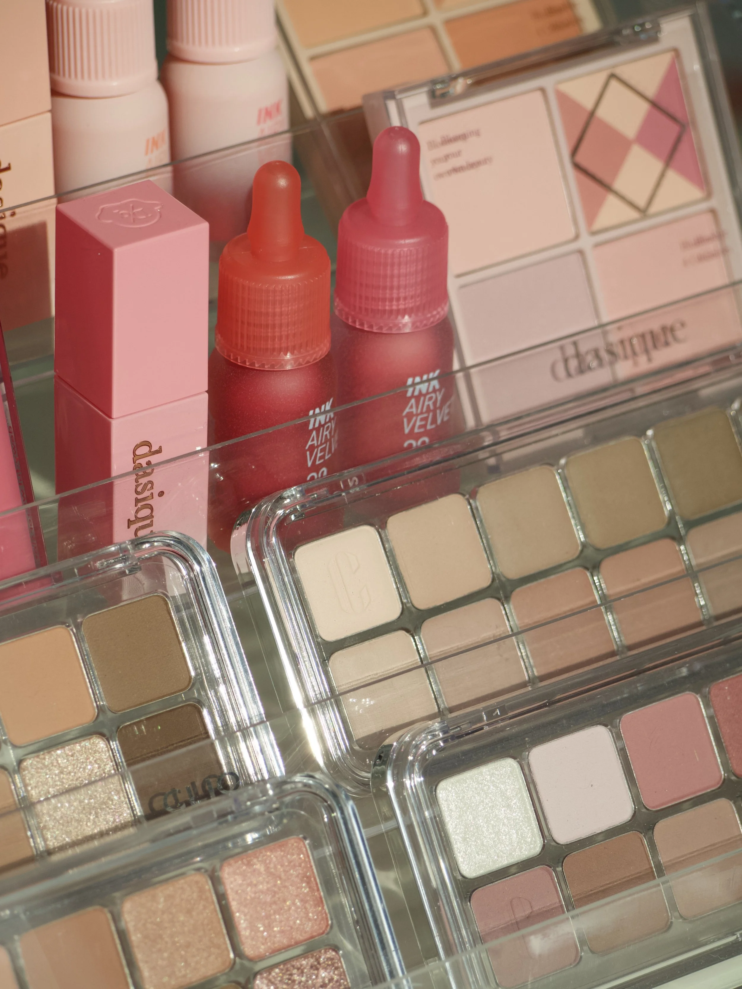 Close-up of a pink cosmetic makeup palette with various eyeshadows, two bottles of pink lip gloss or liquid makeup, and other makeup products in the background.