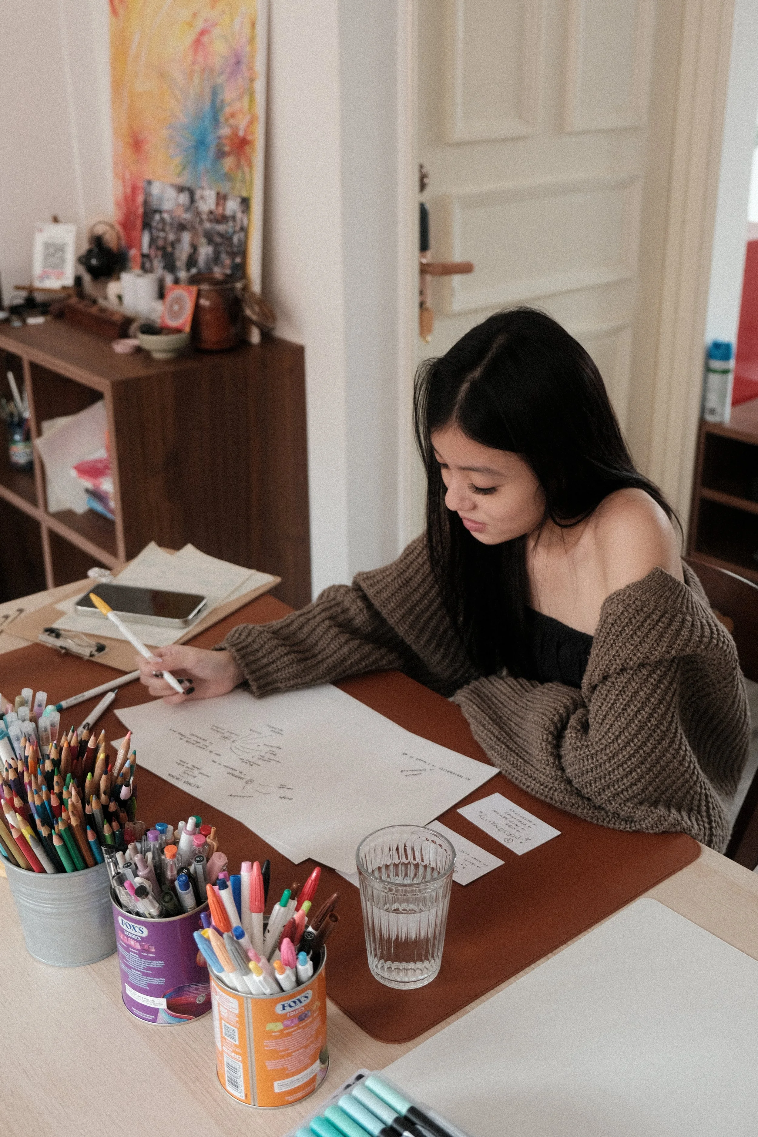 A young woman with long dark hair wearing a brown sweater and a black top, sitting at a desk with art supplies, writing or drawing on large sheets of paper with colored markers. There is a glass of water on the desk, and a smartphone nearby. In the background, there is a wooden bookshelf and other household items.