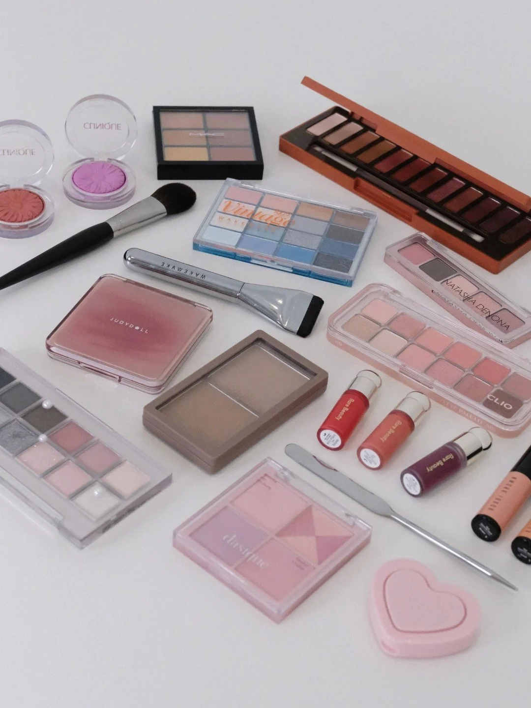 Collection of makeup products including eyeshadow palettes, blushes, lip products, and brushes on a white surface.