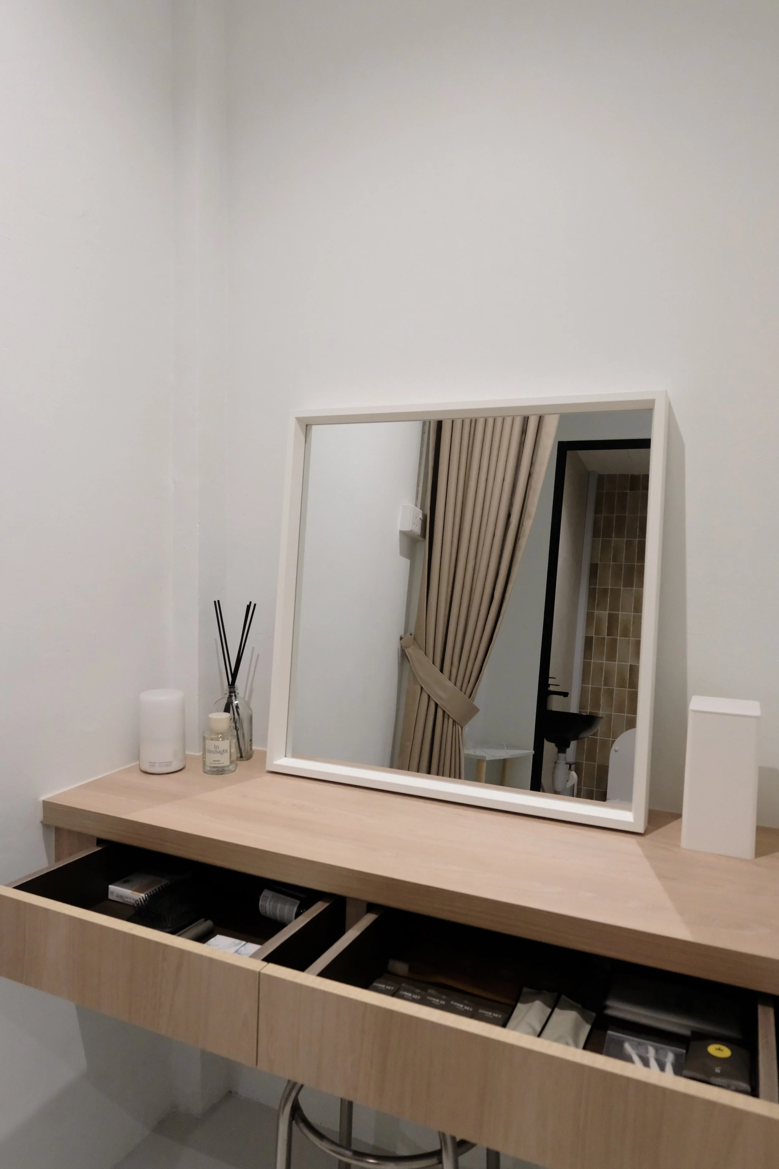 A wooden vanity table with an open drawer. On top, there is a white diffuser, a glass bottle with black sticks, a candle jar, and a white rectangular box. A square white mirror is leaning against the wall, reflecting beige curtains and a partial view of a bathroom with beige tiles and a Korean-style toilet.
