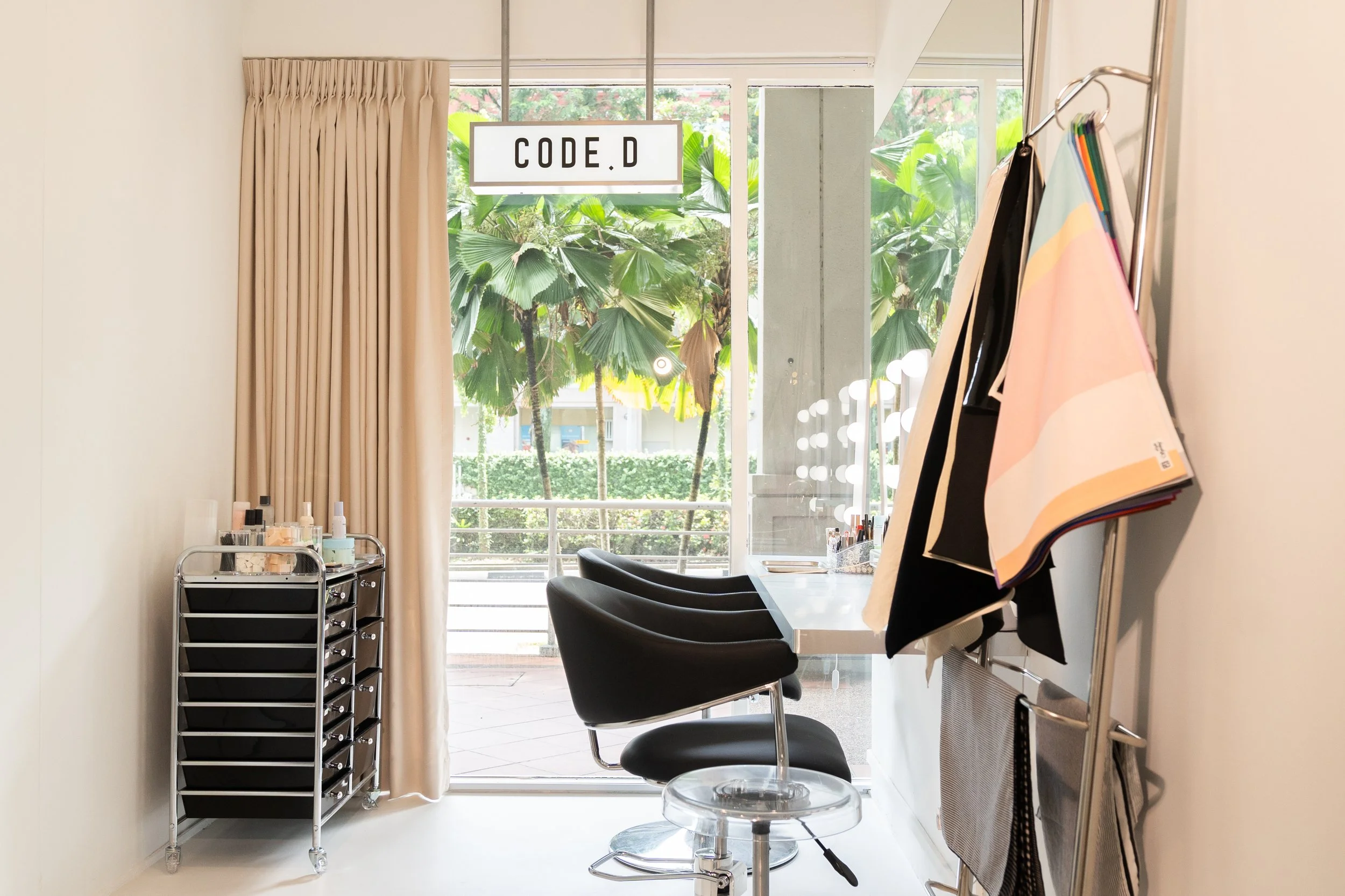 Beauty salon with black styling chairs, a window with beige curtains, a sign that reads 'CODE D', and a metal rack with nail polish bottles, with greenery visible outside.