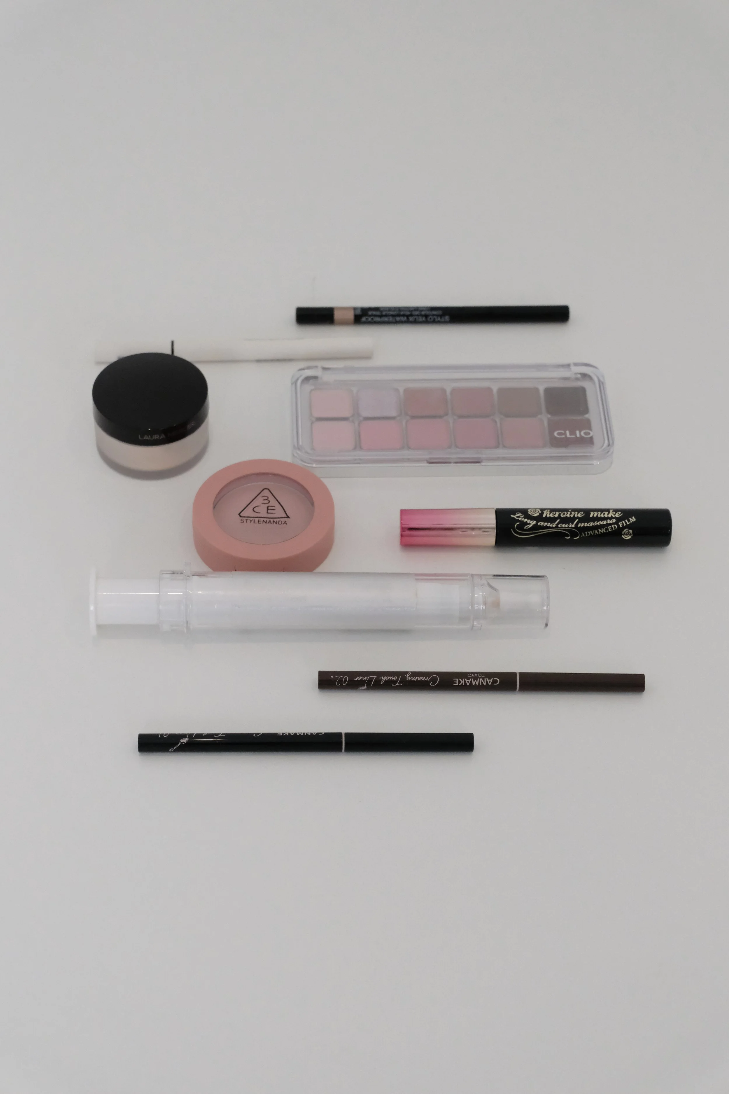 Arrangement of makeup and beauty products on a white surface, including eyeshadow palettes, mascara, eyeliner pencils, a blush, and a lip gloss.