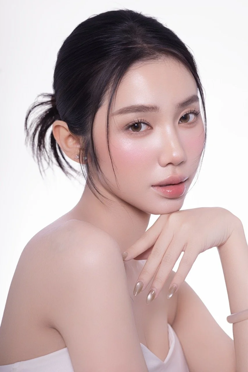 Close-up of a young woman with fair skin, dark hair tied back, light makeup with glossy lips, and a soft expression, resting her chin on her hand against a plain white background.