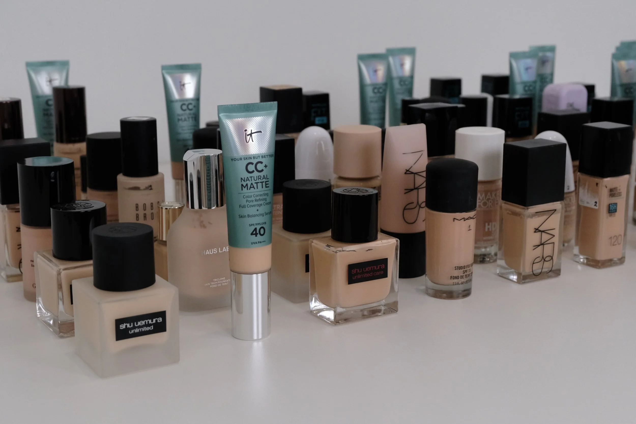 Assorted bottles of foundation and concealer on a white surface, including brands like Shu Uemura, NARS, MAC, Bobbi Brown, and Haus Laboratories.