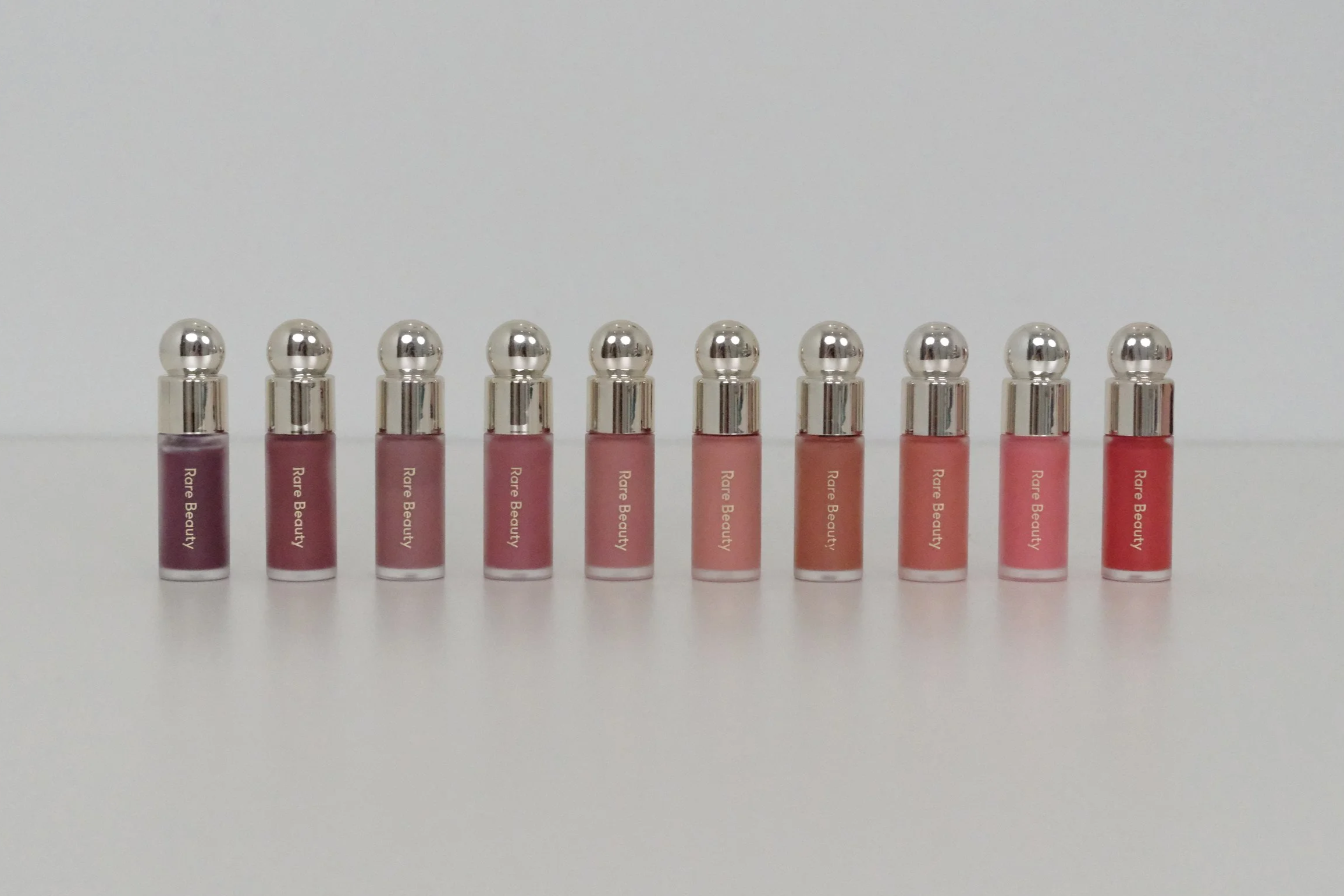Ten lip glosses with silver caps arranged in a row, labeled 'Rare Beauty,' on a white surface with a white background.