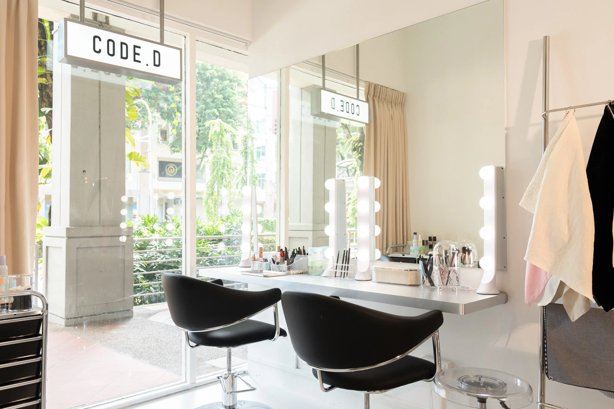 A makeup vanity with bright lights, brushes, beauty products, and two black chairs, positioned in front of a large mirror outside a window with a sign that reads 'CODE D' and 'CODE D' mirrored.