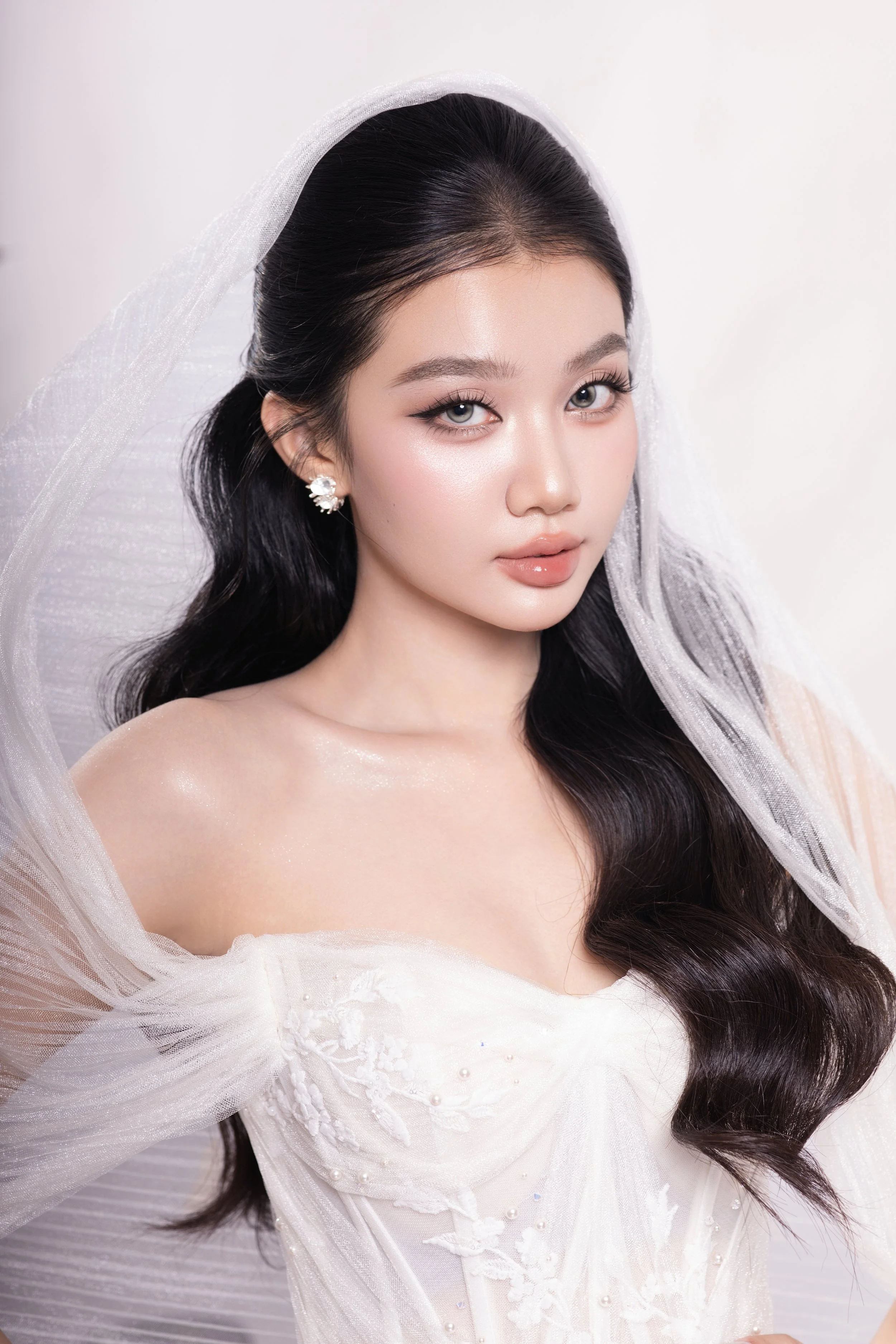 A young woman with long, dark, wavy hair, wearing a white wedding dress and sheer veil, looking at the camera with a neutral expression.
