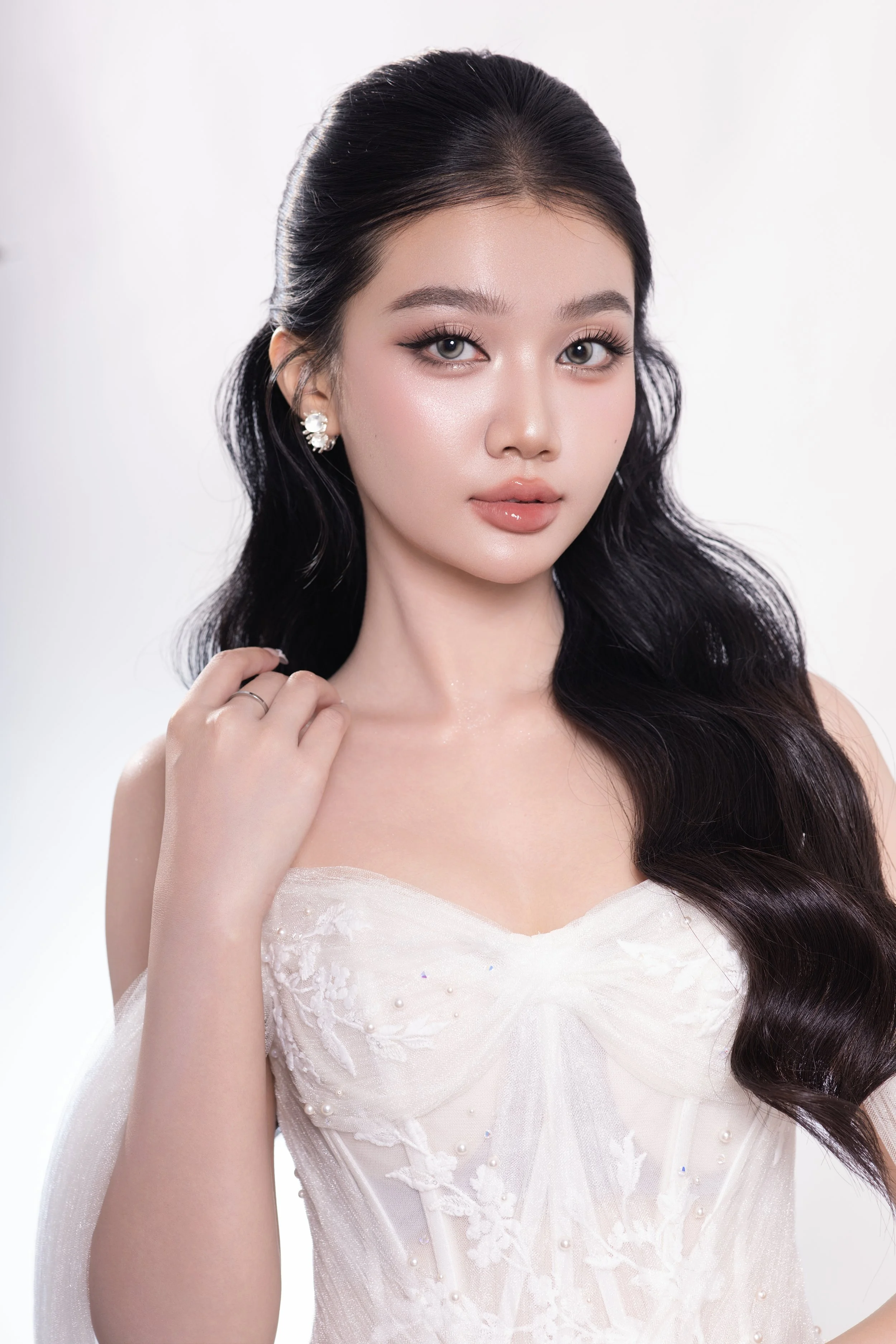 A young woman with long, wavy black hair, wearing elegant earrings and an off-the-shoulder white dress, posing confidently.