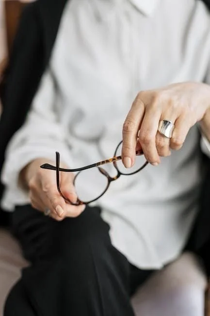 A person holding a pair of eyeglasses with both hands, wearing a white shirt and a large ring on their right ring finger.