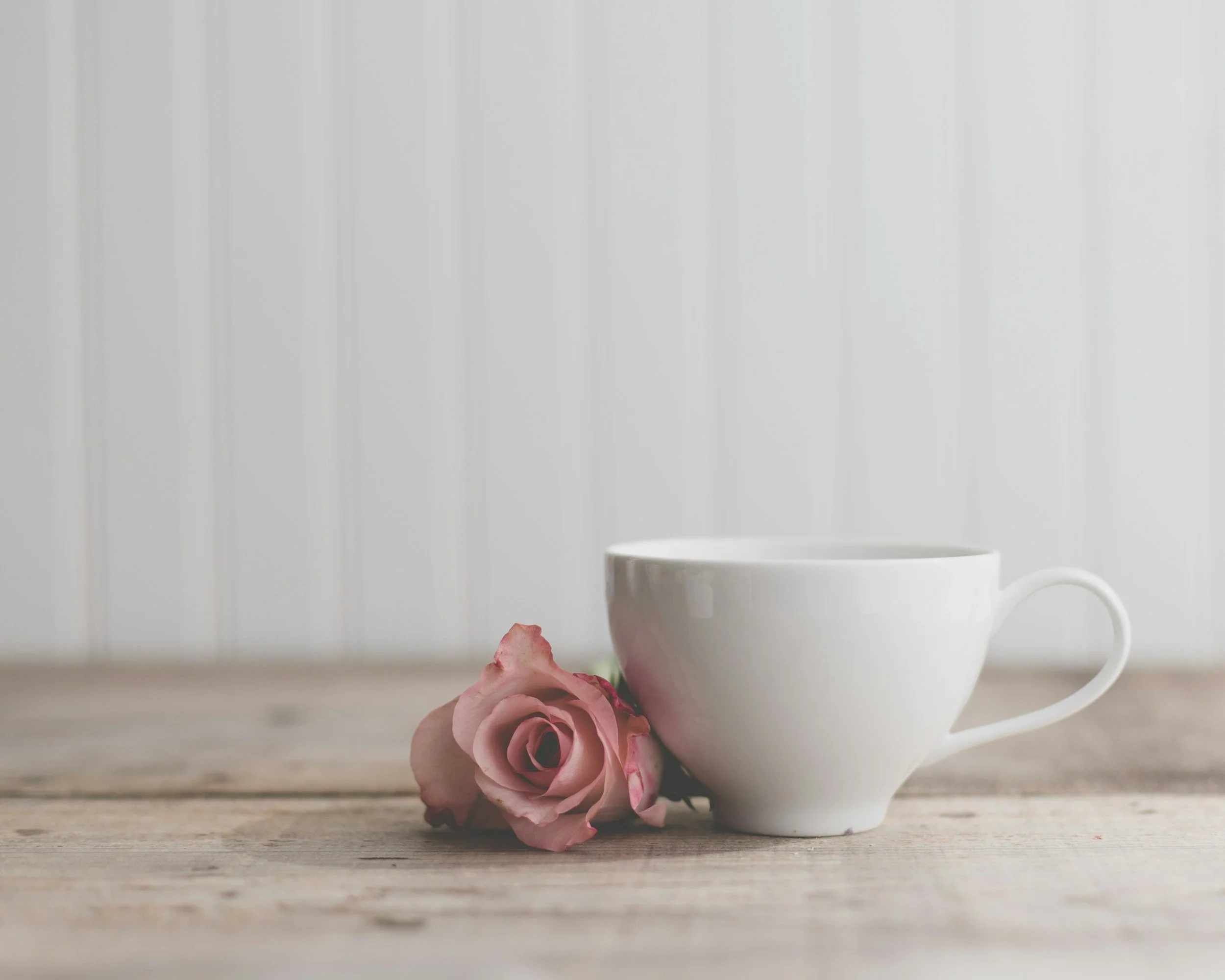 A white ceramic coffee cup next to a pink rose on a wooden surface with a white paneled wall in the background.