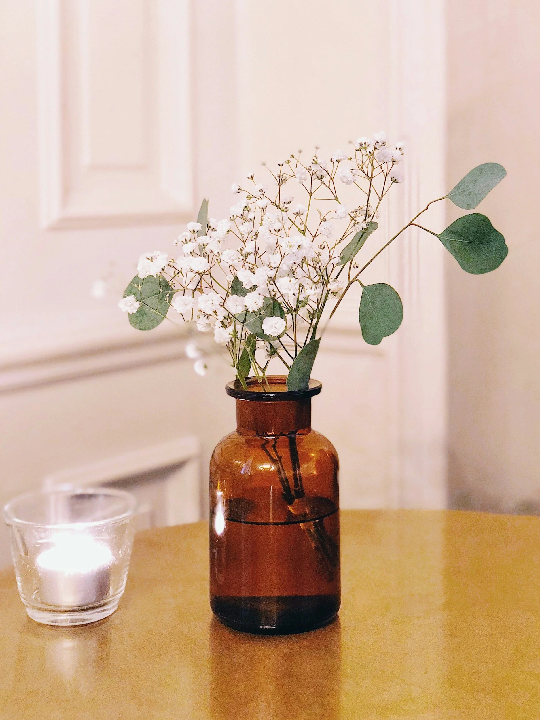 A brown glass vase with white flowers and green leaves on a wooden surface, with a lit candle in a glass holder nearby.