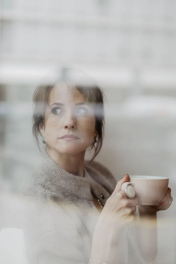 A woman with short brown hair and earrings, sitting indoors and looking out the window while holding a cup.