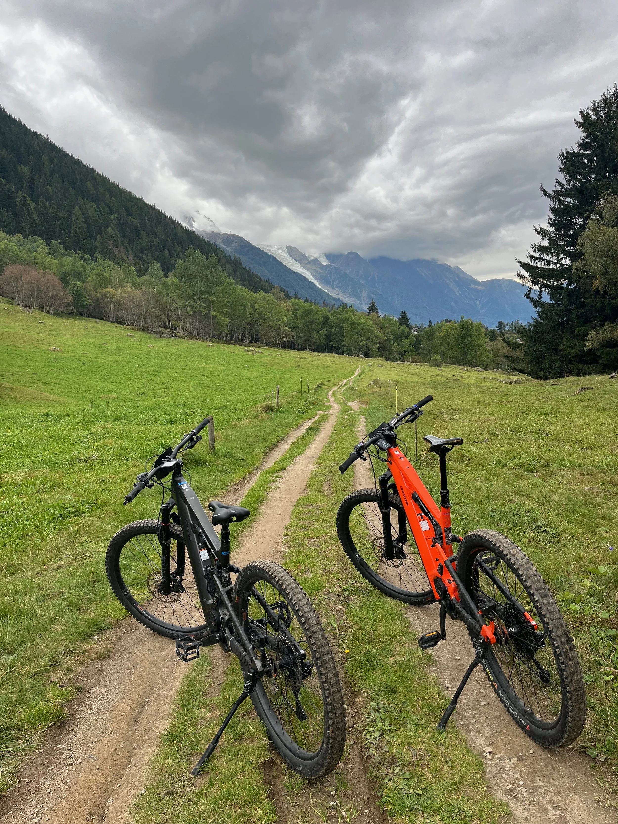 Deux vélos électriques sur un sentier de montagne avec des montagnes et un ciel nuageux en arrière-plan.