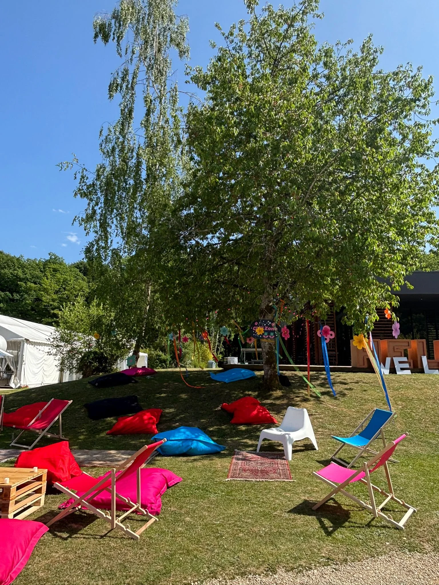 Décor en plein air avec des chaises pliantes colorées, des coussins rouges et noirs, un arbre décoré avec des guirlandes et des fleurs en plastique, sous un ciel bleu, en arrière-plan un bâtiment en bois et un panneau "LOVE".
