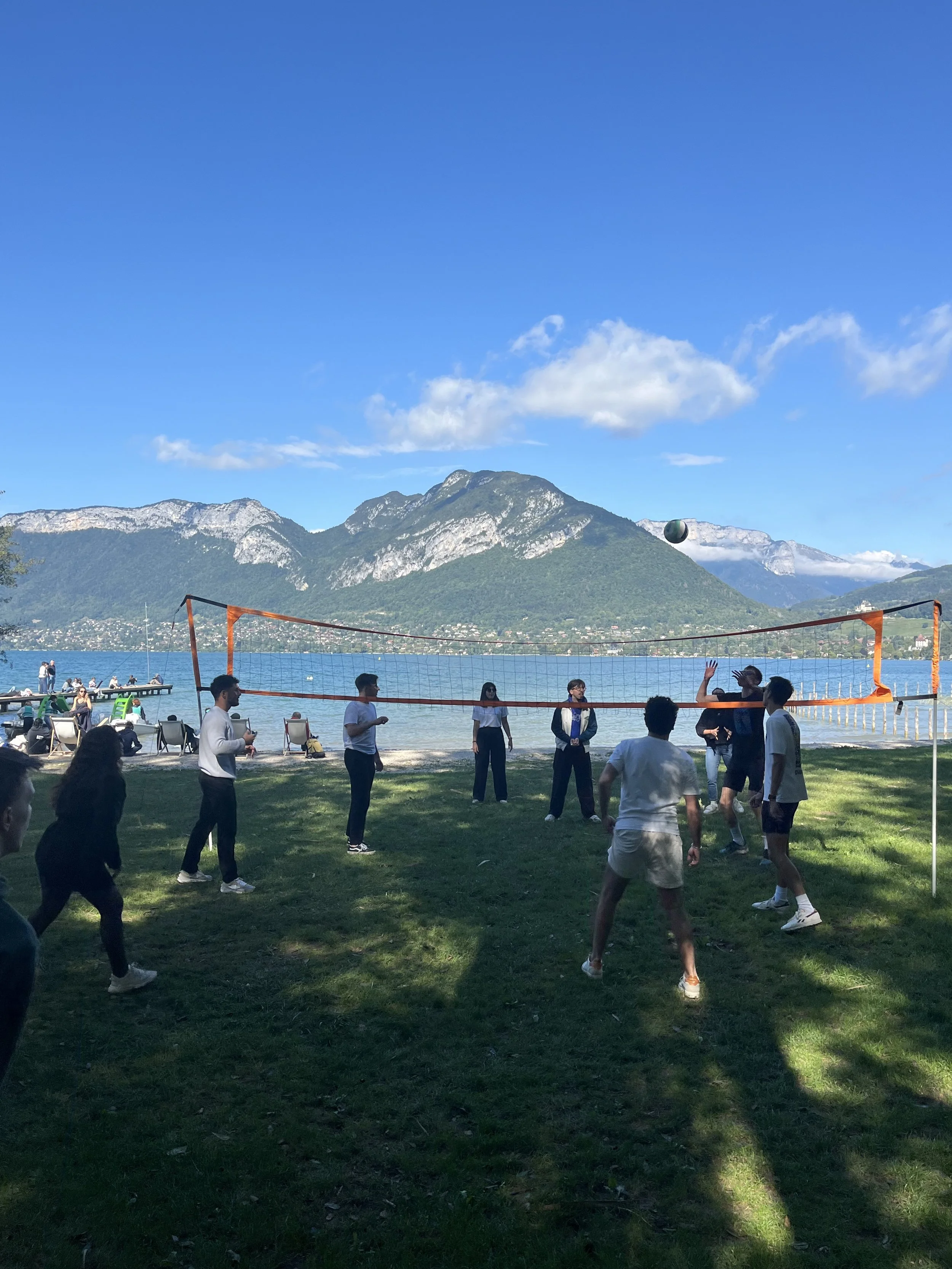 Groupe de personnes jouant au volleyball sur une plage avec vue sur un lac et des montagnes en arrière-plan, sous un ciel bleu.