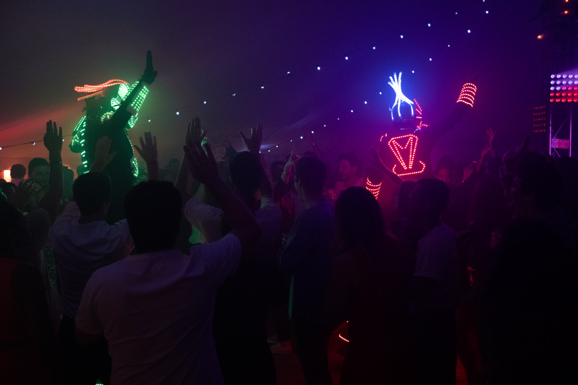Fête nocturne avec des danseurs en costumes lumineux LED, foule de personnes dansant sous des lumières colorées.
