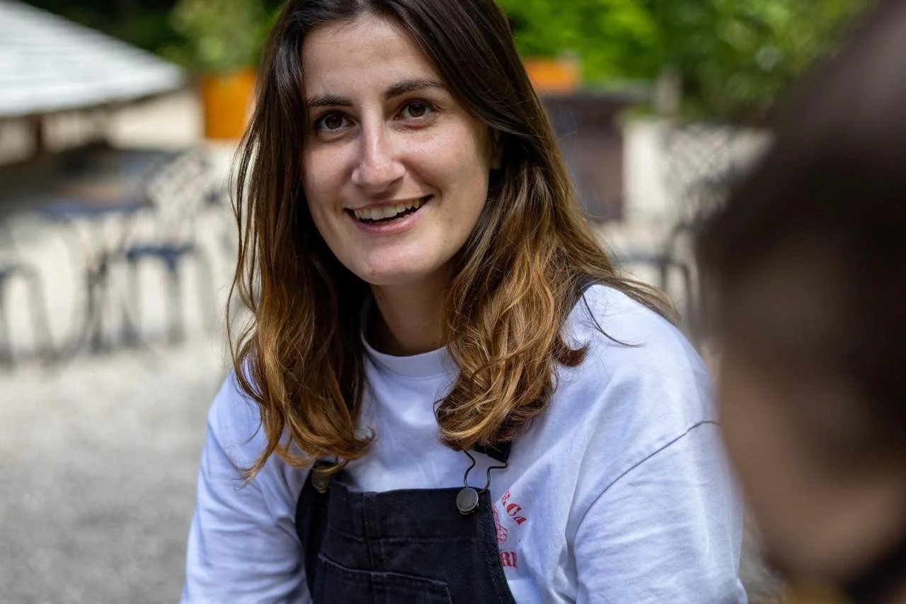Une femme avec des cheveux bruns, portant un T-shirt blanc et un tablier noir, sourit à l'extérieur, en arrière-plan des chaises et une table