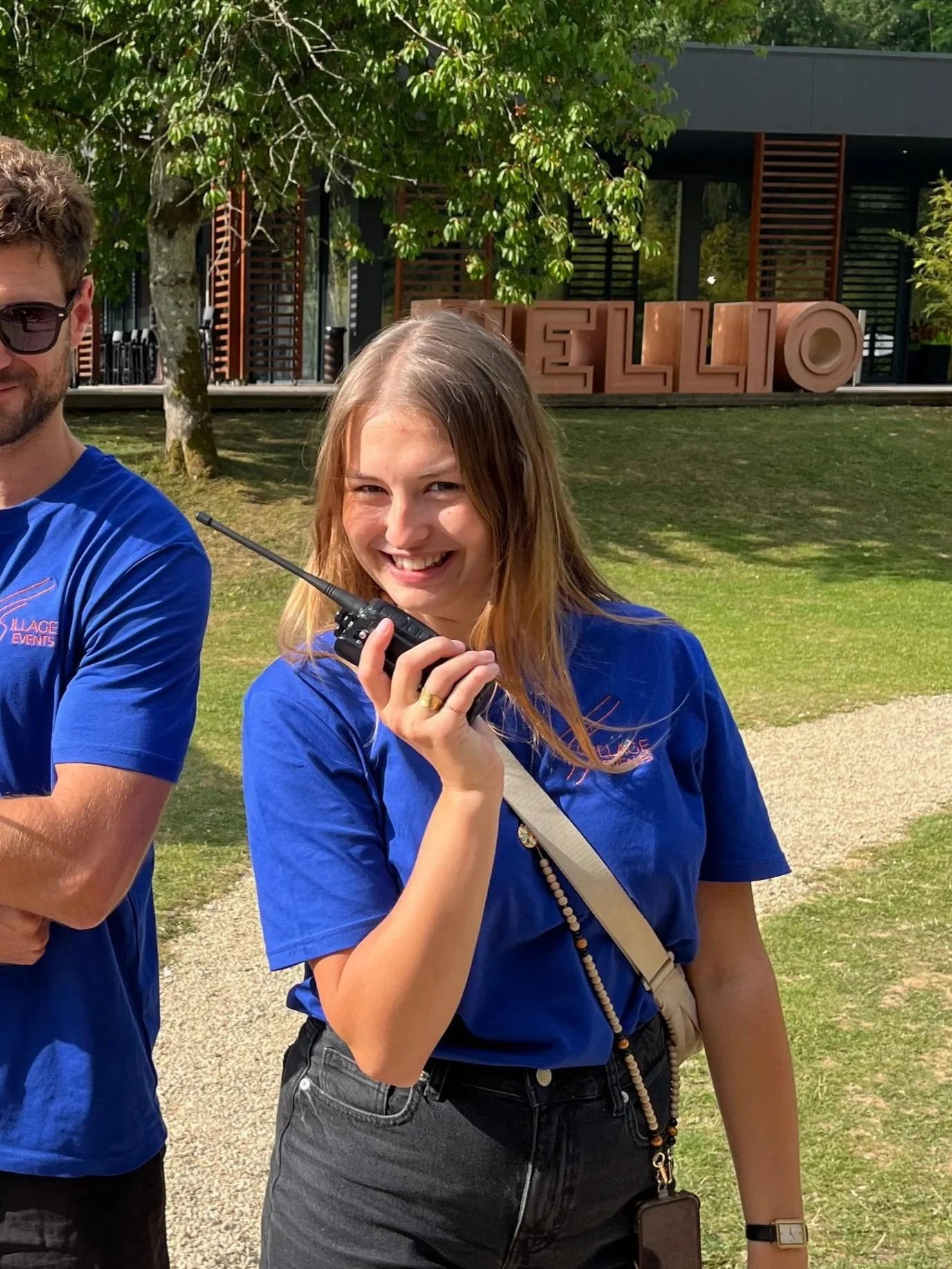 Une jeune femme souriante tenant une radio en main, en plein air avec des arbres et un bâtiment en arrière-plan, portant un t-shirt bleu et un sac en bandoulière, lors d'un événement.