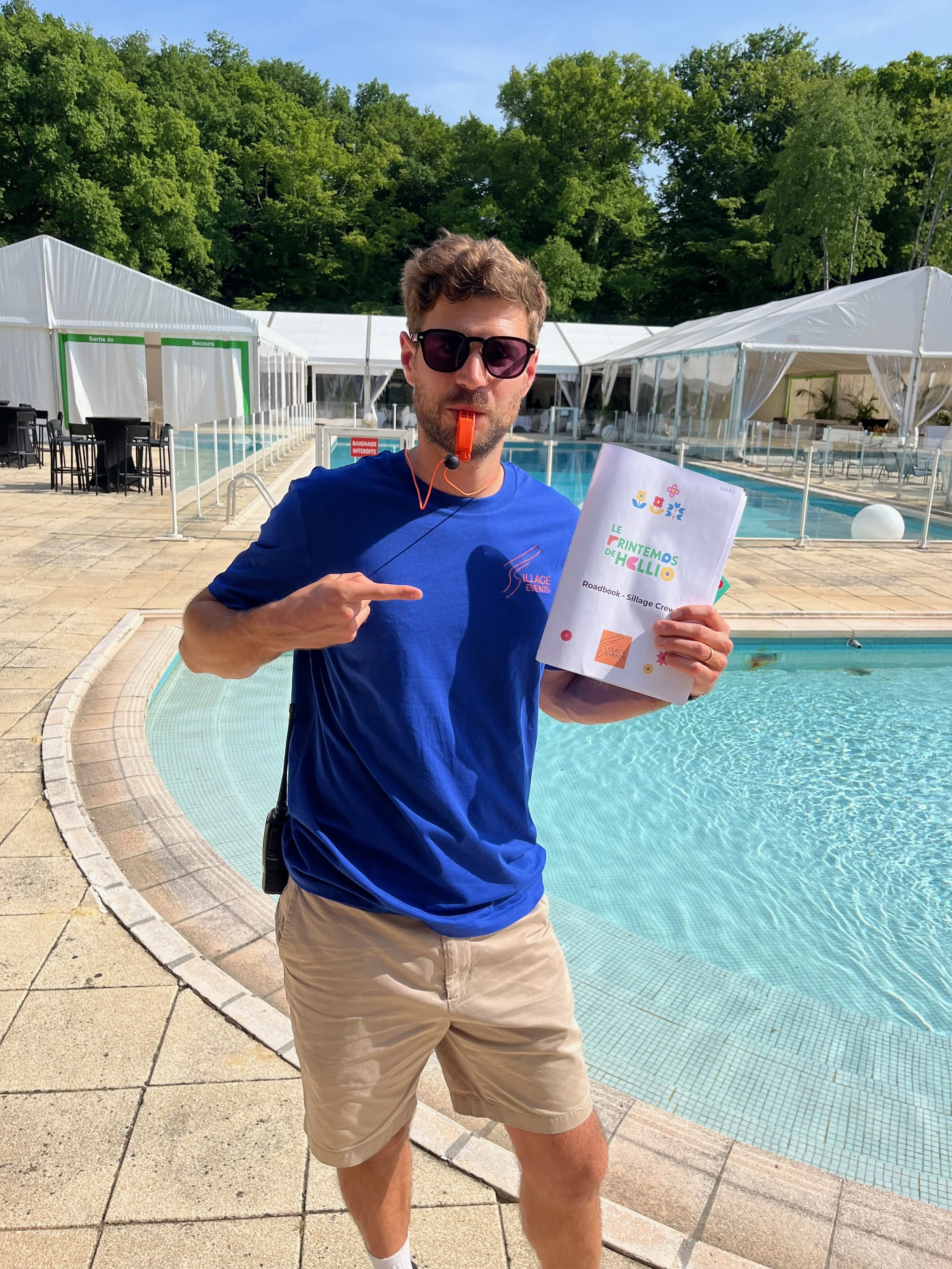 Un homme portant des lunettes de soleil, une t-shirt bleu, et un short beige, se tient près d'une piscine en pointant vers un document qu'il tient dans la main. La scène se déroule dans un espace en plein air avec des tentes blanches en arrière-plan, entouré d'arbres verts.
