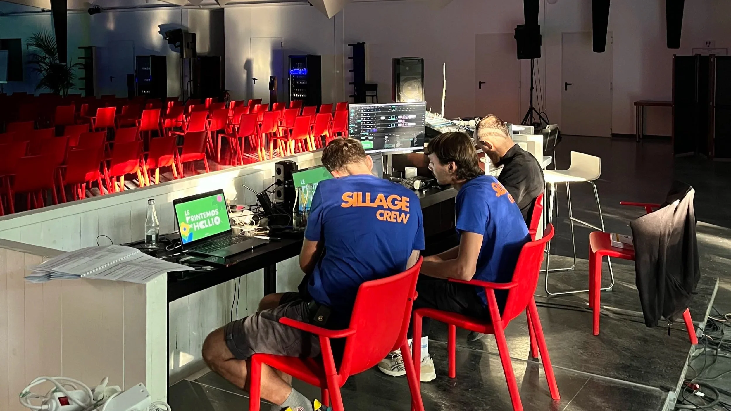 Trois techniciens ou DJ travaillent à une table de contrôle avec des écrans et du matériel audio dans une salle vide, probablement une salle de spectacle ou de conférence, avec des chaises rouges alignées au fond.