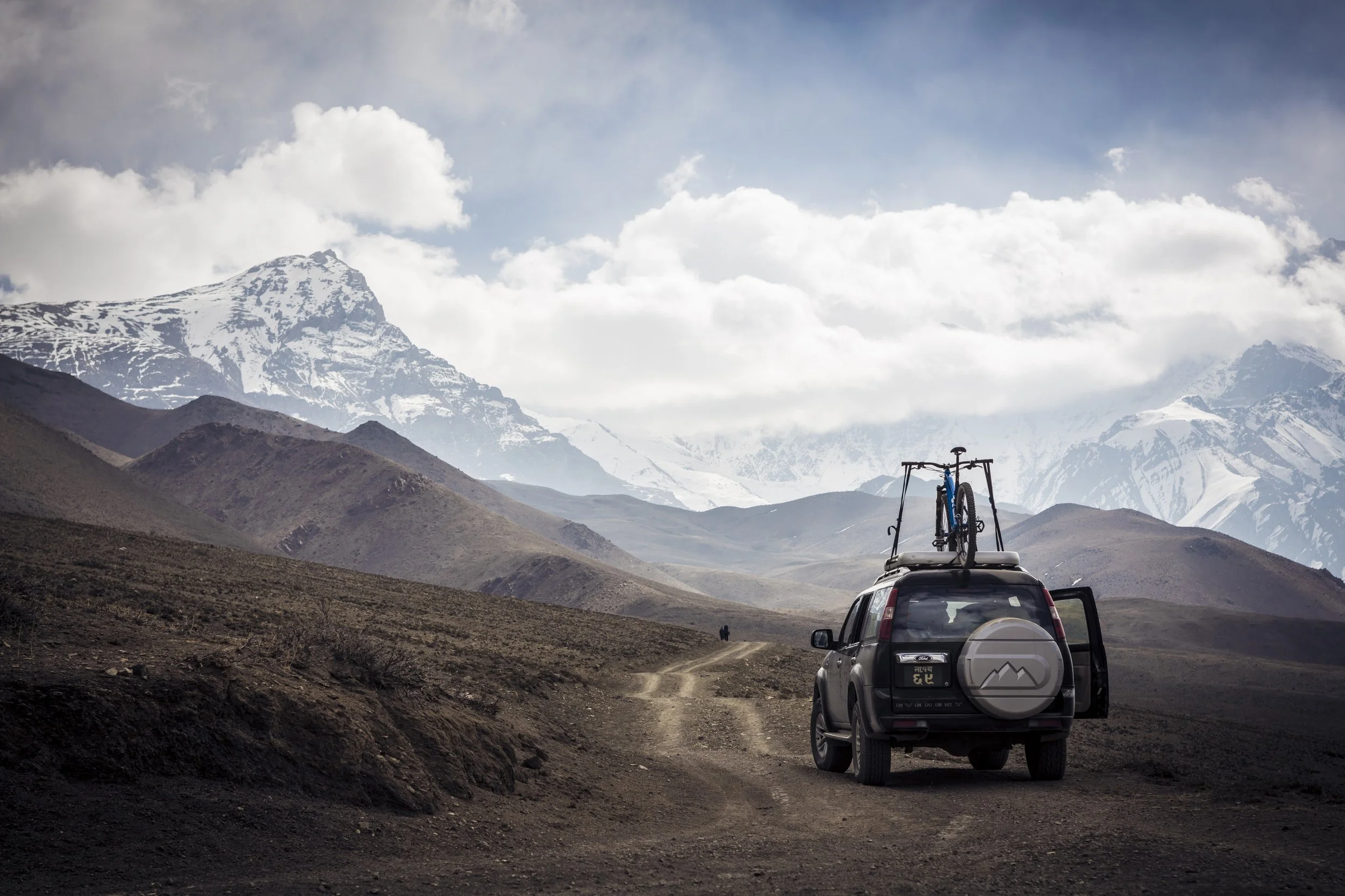 A black SUV with a bicycle mounted on the roof and a spare tire on the back, parked on a dirt road in a mountainous landscape with snow-capped peaks and cloudy skies.