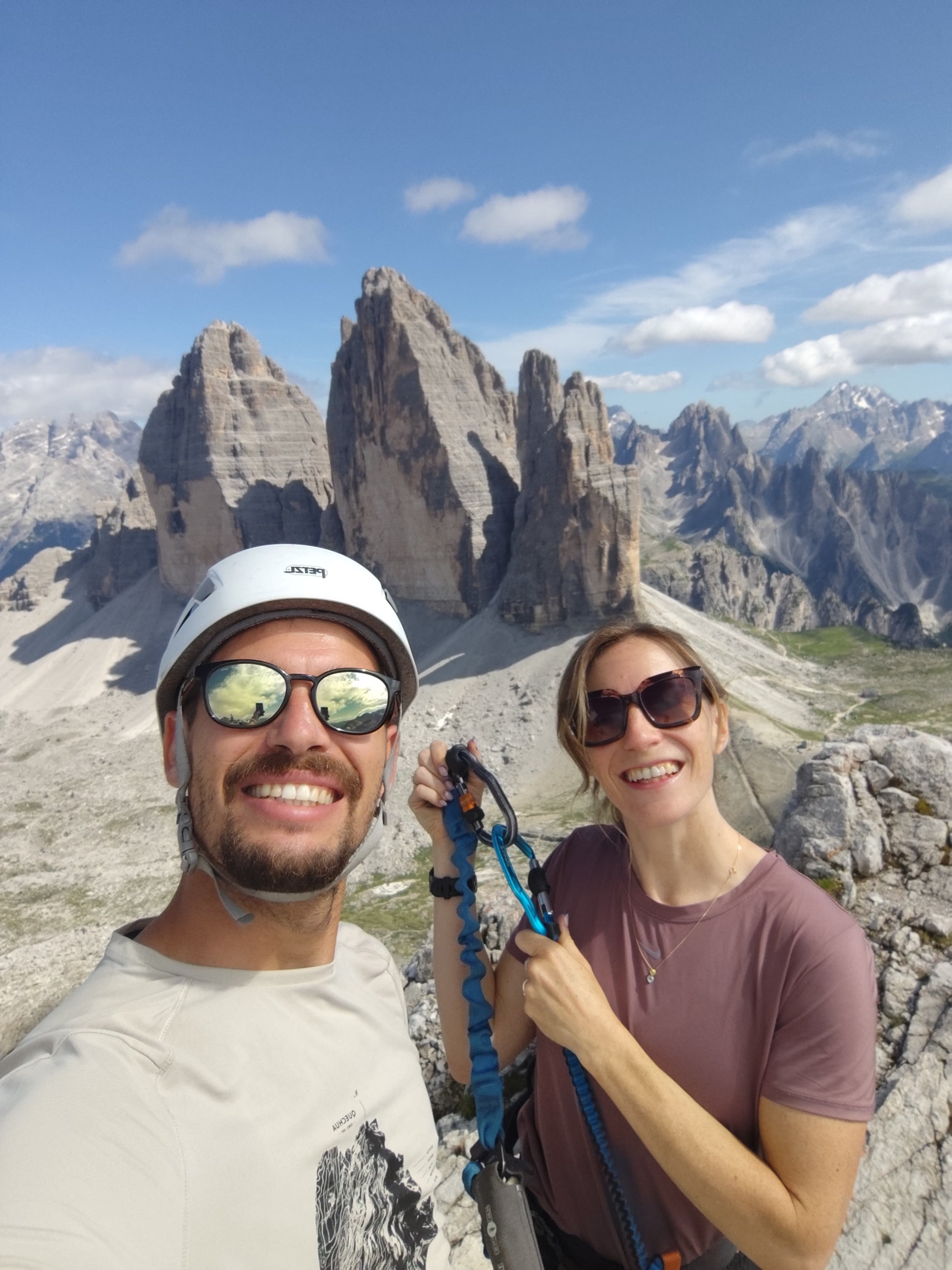 Smiling man wearing a helmet and sunglasses and woman with sunglasses, holding climbing gear, in front of mountain peaks in a rocky landscape on a bright, partly cloudy day.