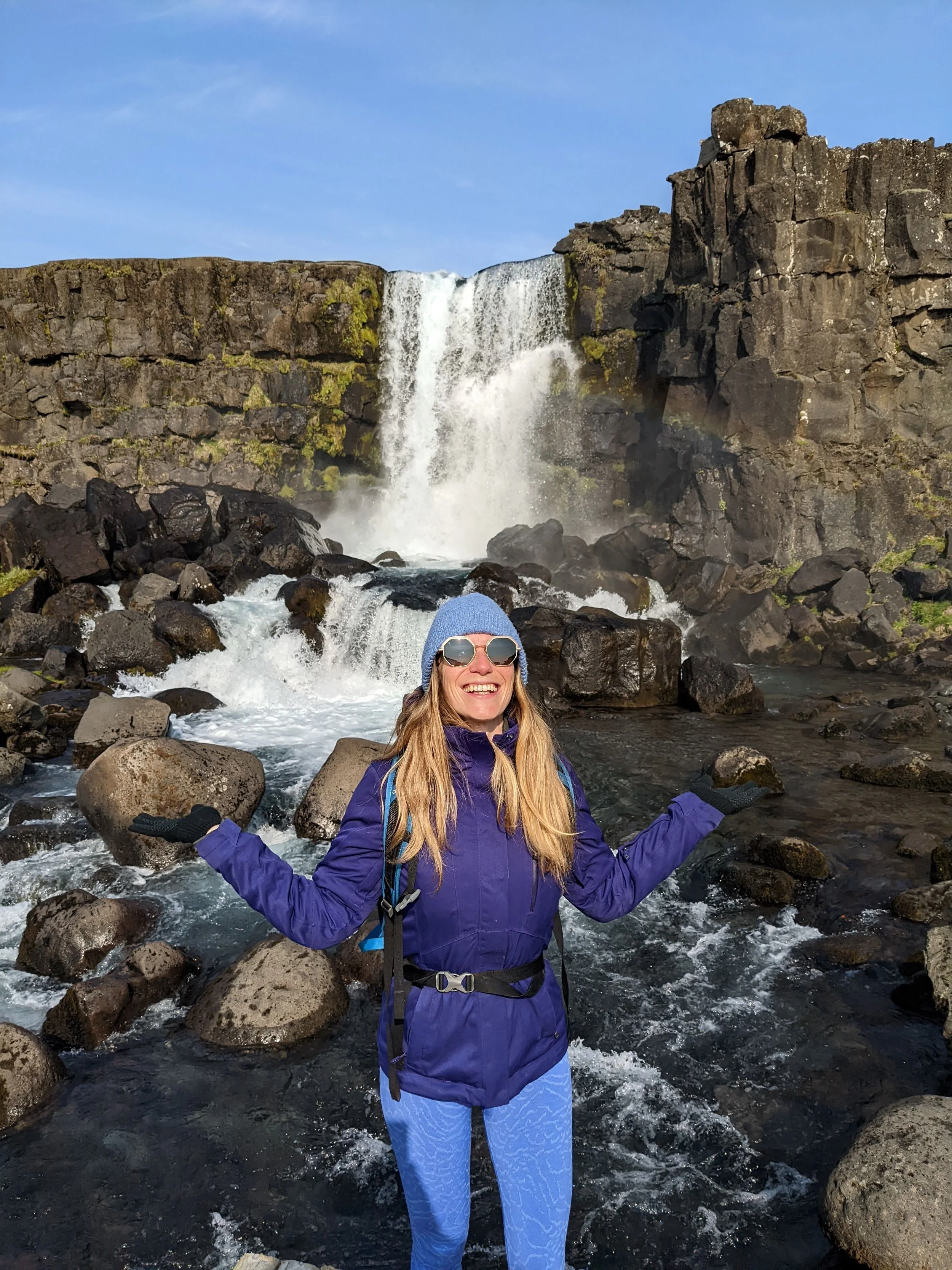 A woman smiling with arms outstretched standing in a rocky river in front of a large waterfall, dressed in outdoor gear including a purple jacket, blue beanie, sunglasses, and gloves.