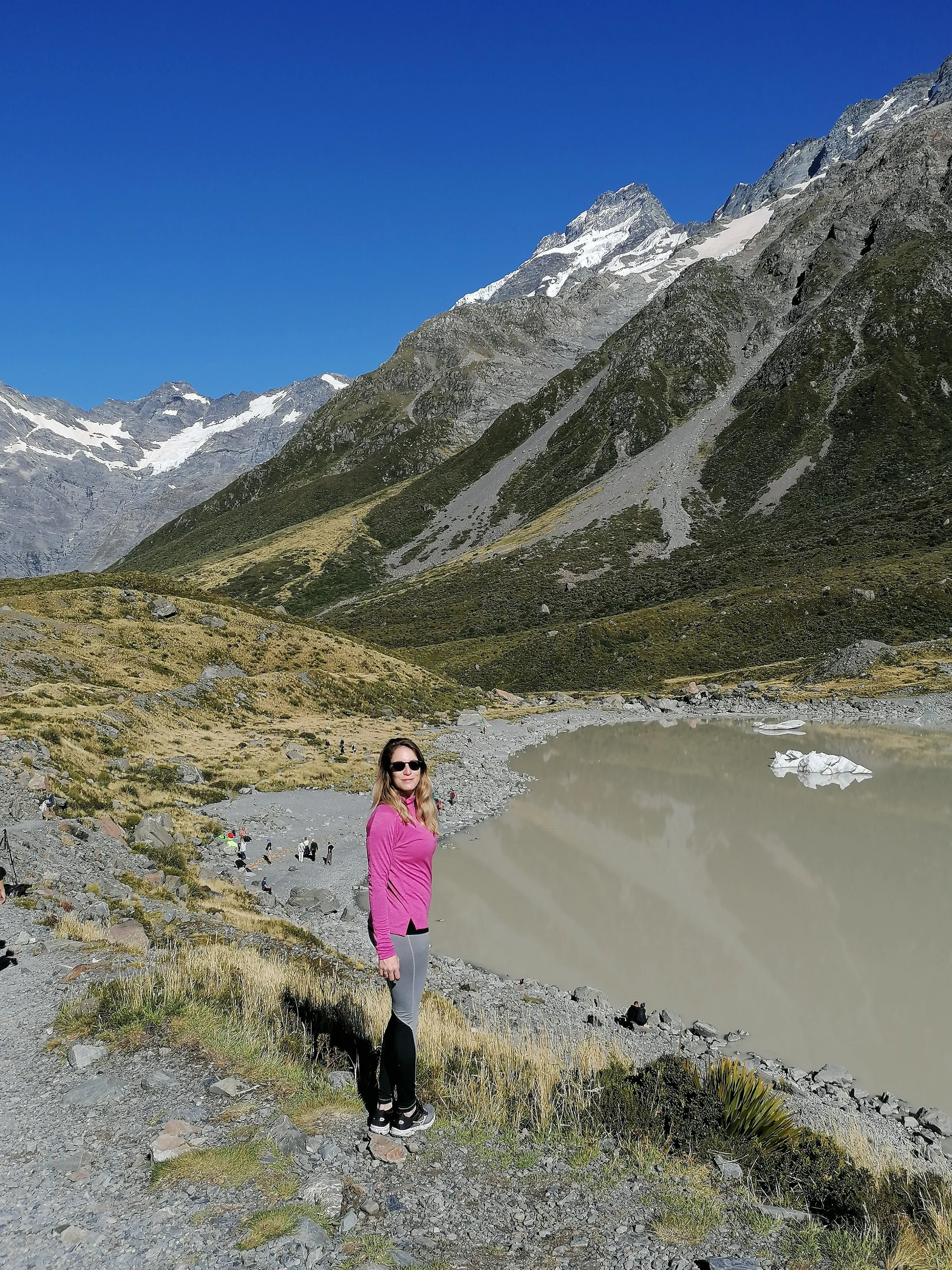 A woman in pink and gray outdoor clothing standing on rocky ground near a lake in a mountainous landscape with snow-capped peaks in the background.