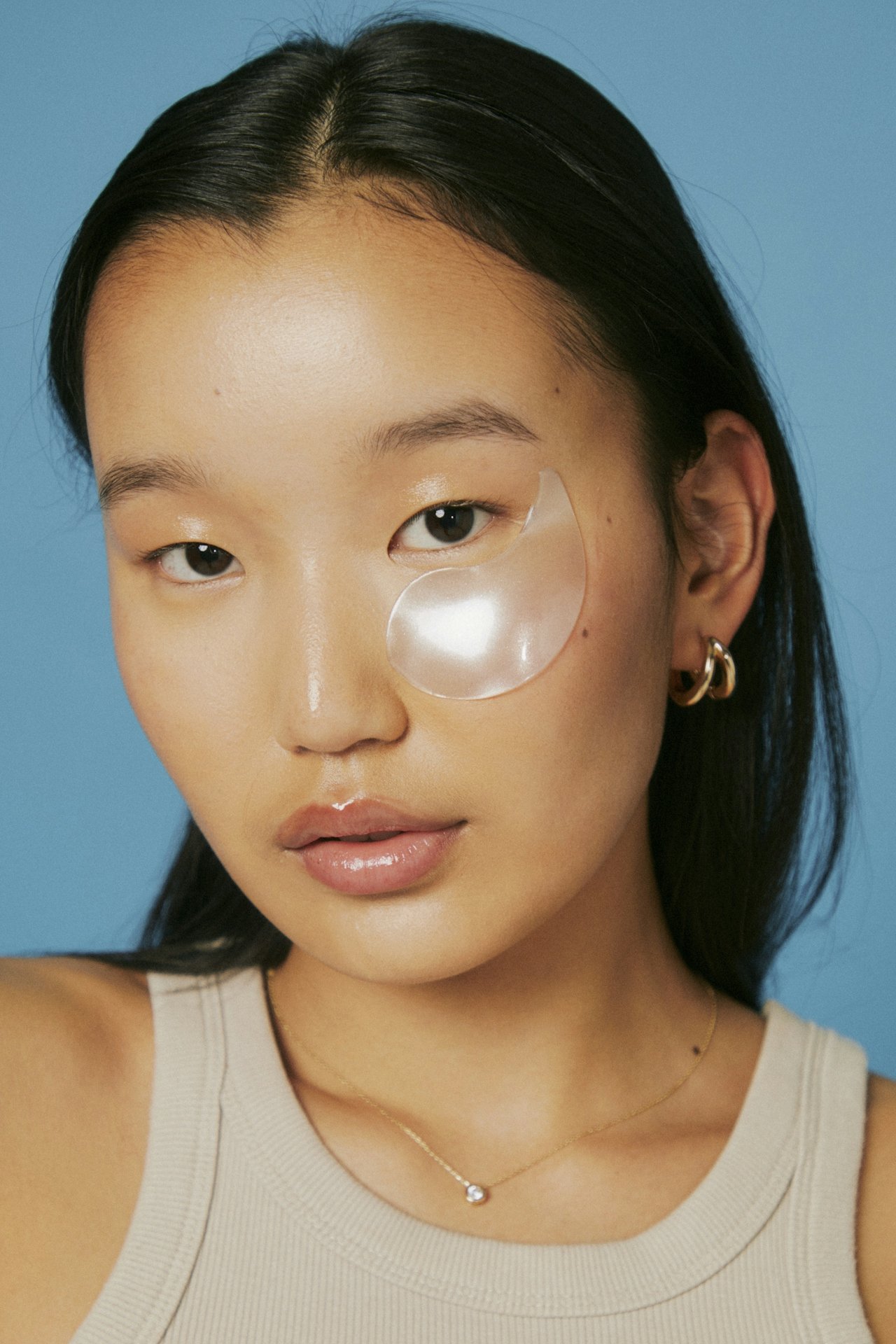 A young woman with dark hair wearing a beige tank top and gold jewelry, including earrings, a necklace, and a small chain, with a blue background. She has a translucent under-eye gel patch on her right eye.