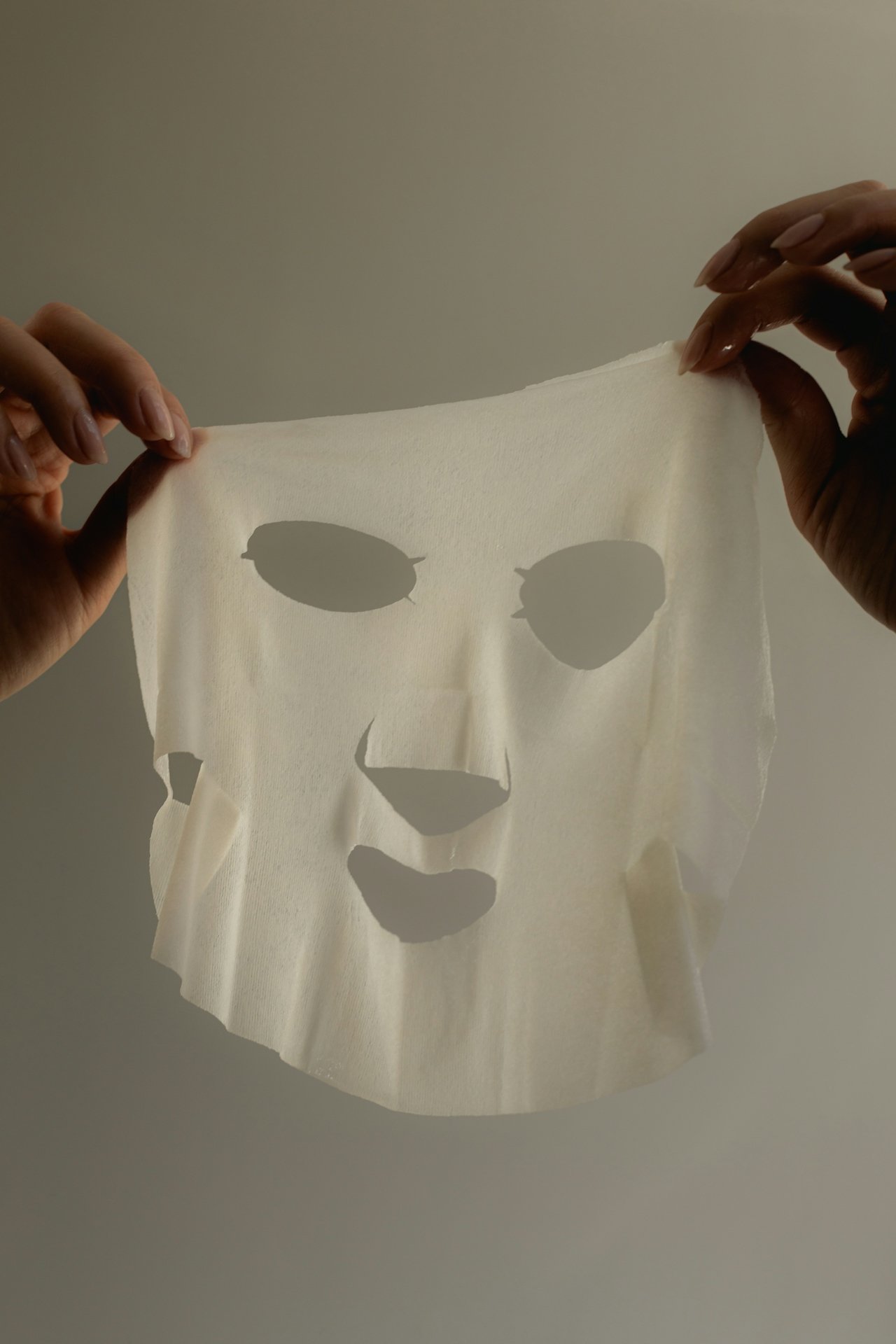 Person holding a white sheet with a face cutout against a plain background.