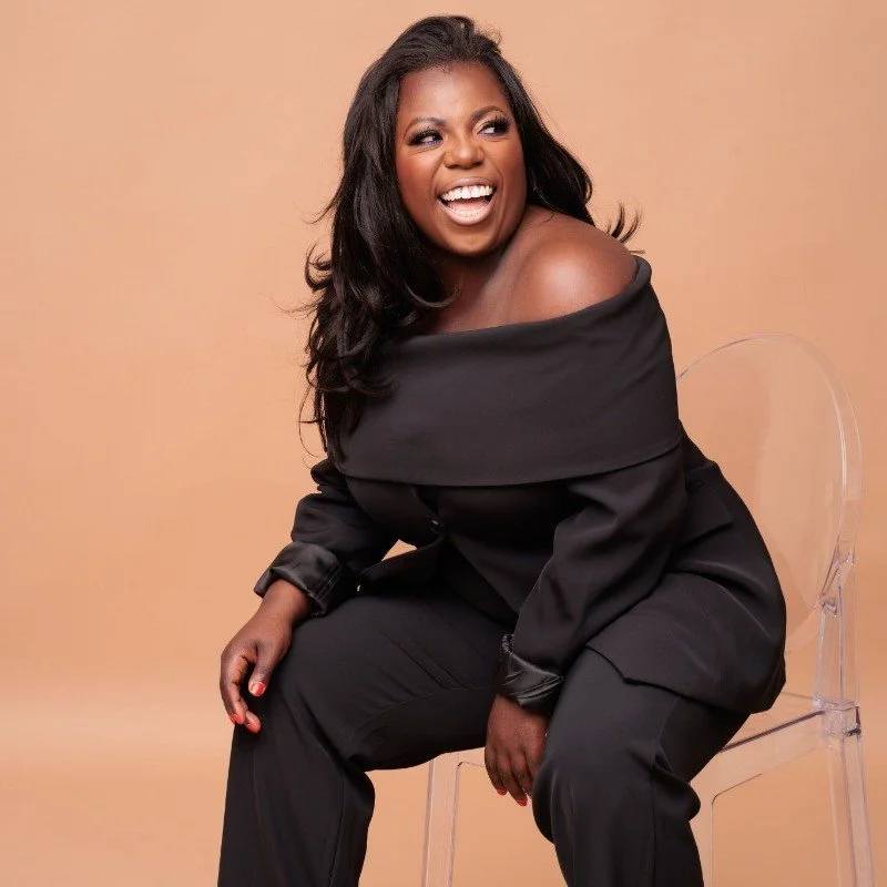 A woman with dark hair and makeup, wearing an off-the-shoulder black outfit, sitting on a clear chair, smiling and looking to her left against a beige background.