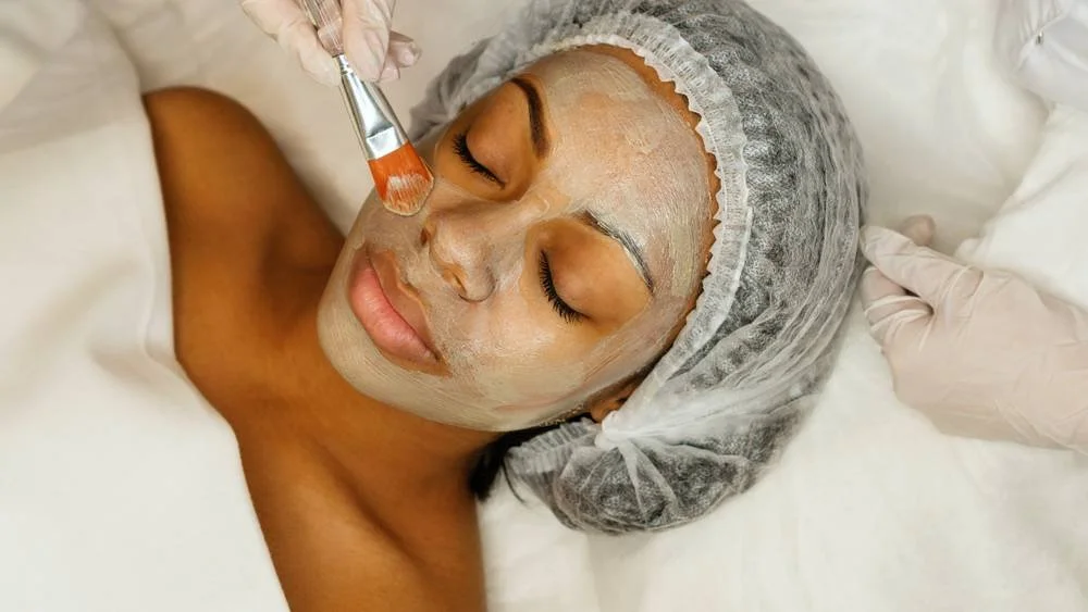 A woman is receiving a facial treatment, with her eyes closed, wearing a hair cap, as a skincare professional applies a mask to her face with a brush.