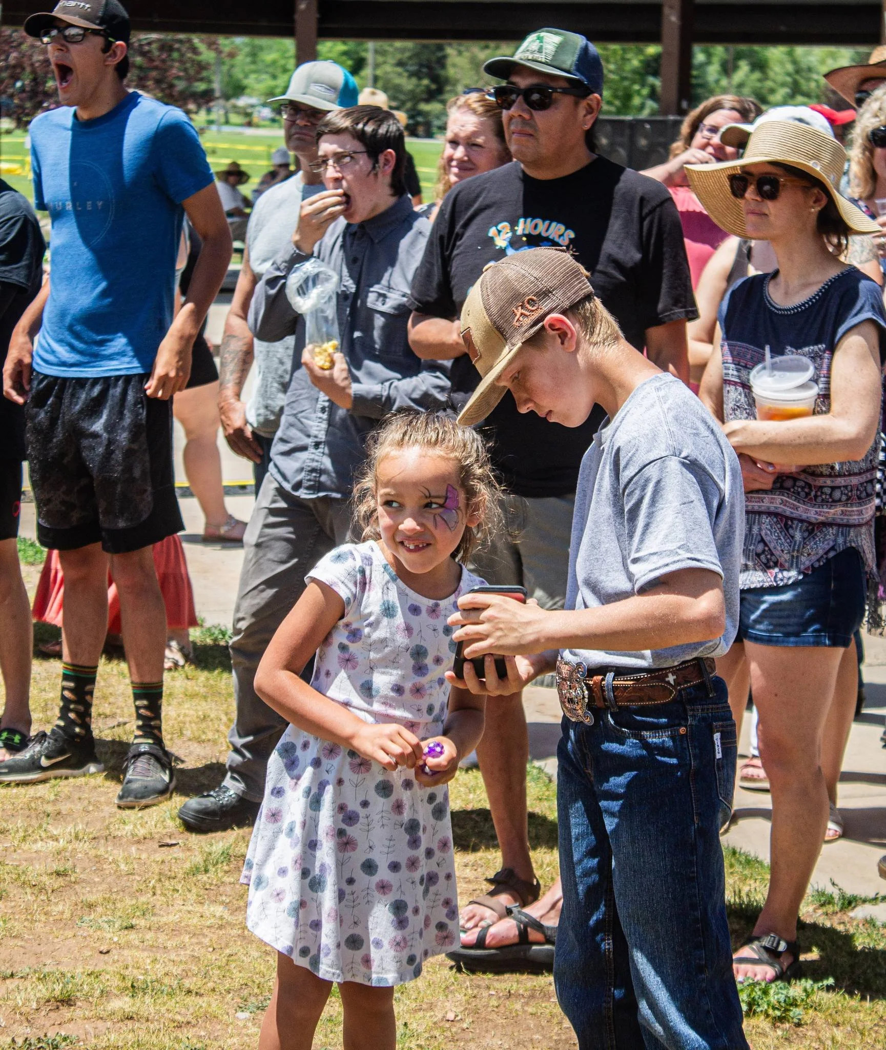 A young girl with face paint and a dress is smiling as she talks to a teenage boy wearing a cap and gray shirt, who is showing her something on his phone, in a crowd outdoors on a sunny day