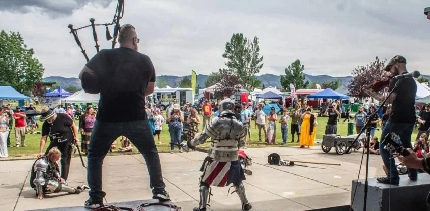 Street performer in a robot costume performing on stage at an outdoor fair, with a musician playing violin to the right and a crowd watching.
