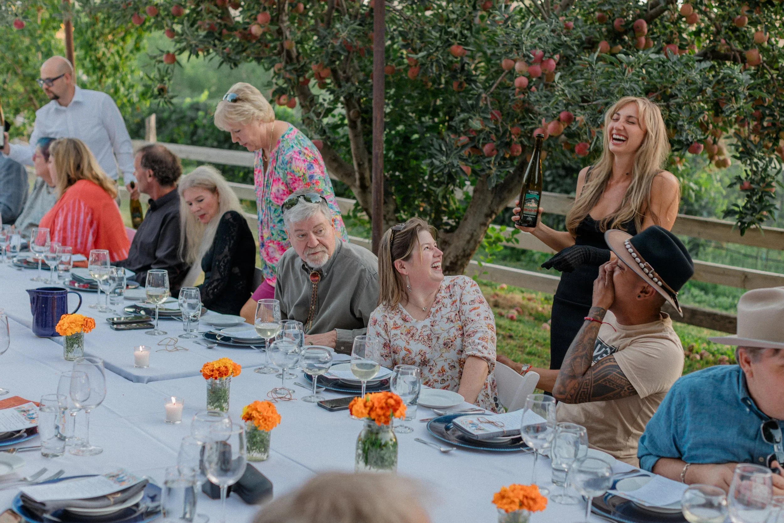People gathered at an outdoor party, sitting at a long table decorated with orange flowers and candles, with a woman in black serving drinks and others engaging and smiling.