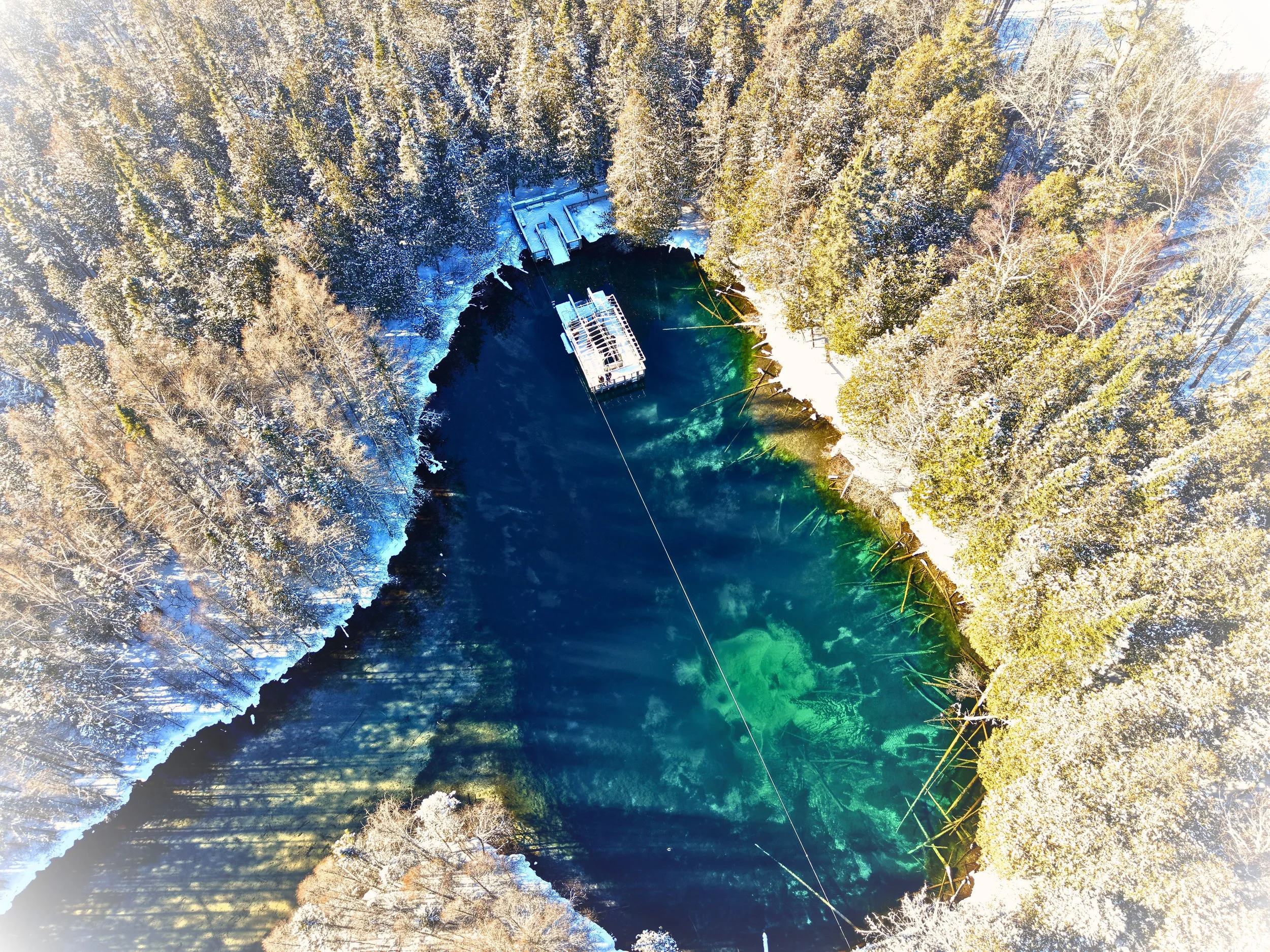 Aerial view of a small lake surrounded by snow-covered trees in winter, with a dock and a boat in the water.
