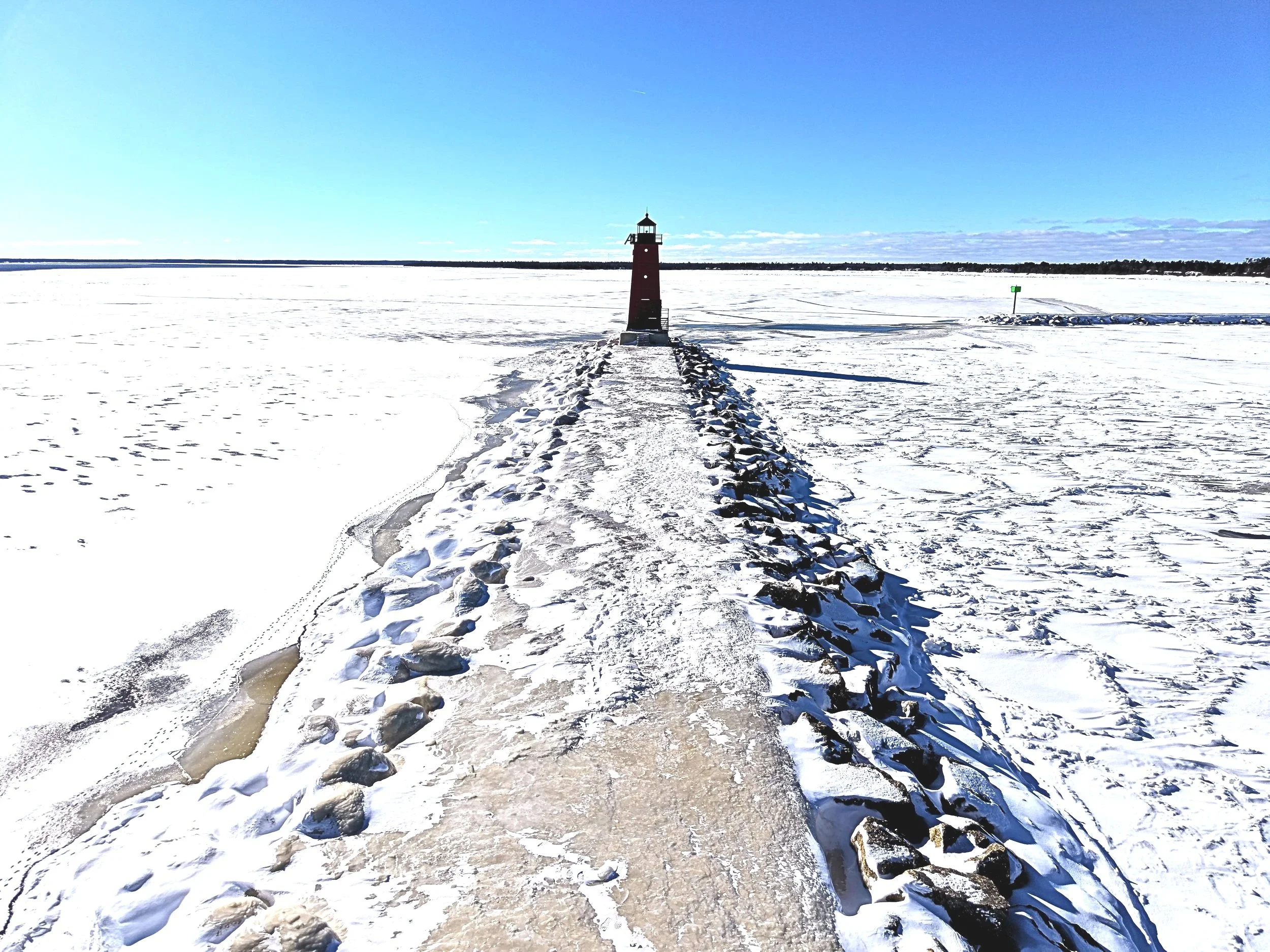 A lighthouse standing on a snow-covered pier extending into a frozen lake under a clear blue sky.