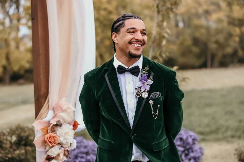 A man in a green velvet suit with a black bow tie, white shirt, and floral boutonniere, smiling outdoors in front of a decorated wedding arch with pink and orange flowers, green foliage, and light-colored fabric.