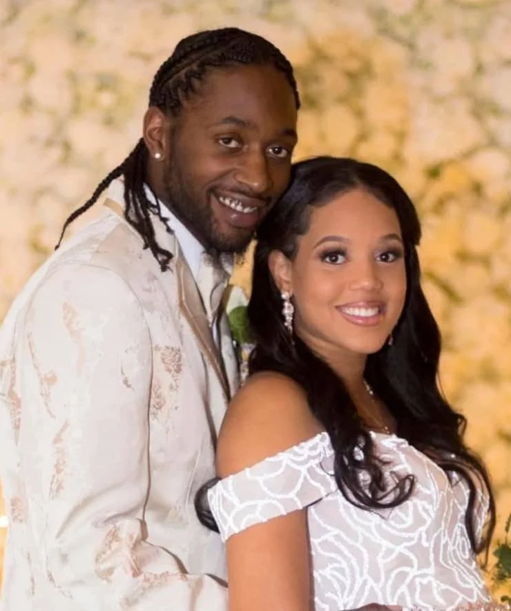 A smiling couple at their wedding, with the man wearing a light-colored patterned suit with long braids and the woman in a white off-the-shoulder dress with dark hair and earrings, in front of a winter wonderland floral background.by Skye High Events