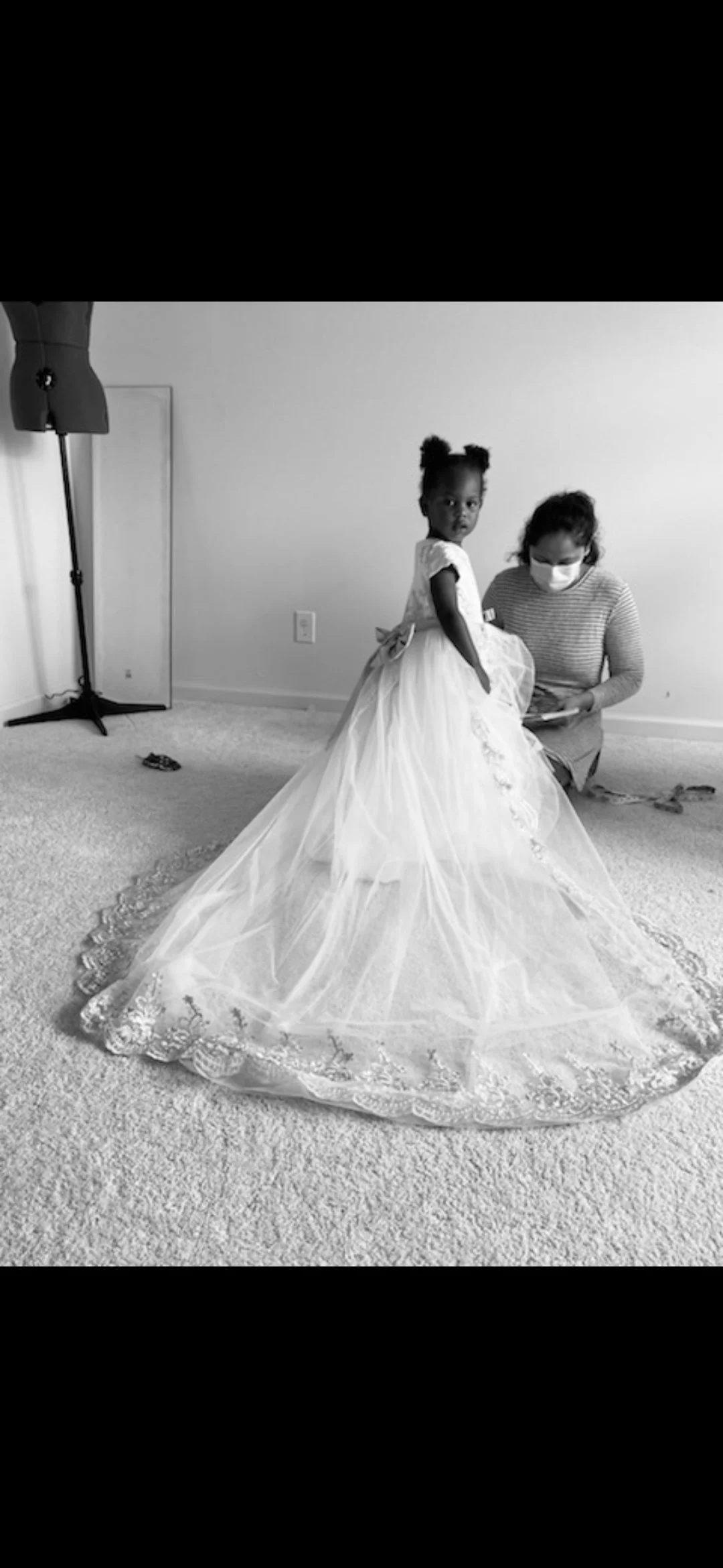 A young girl in a formal dress, standing on a carpeted floor, with a woman kneeling beside her wearing a mask and striped shirt. The background is a plain wall, and a mannequin and some miscellaneous items are visible in the room.