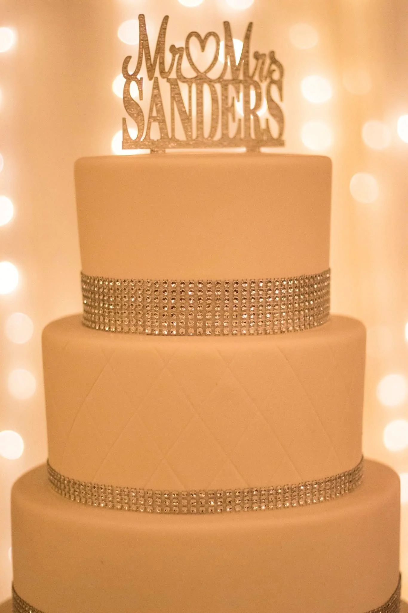 Three-tiered wedding cake with rhinestone bands, topped with a glittery topper reading 'Mom & Dad Sanders' with a heart between 'Mom' and 'Dad,' set against a blurred backdrop of warm, glowing lights.