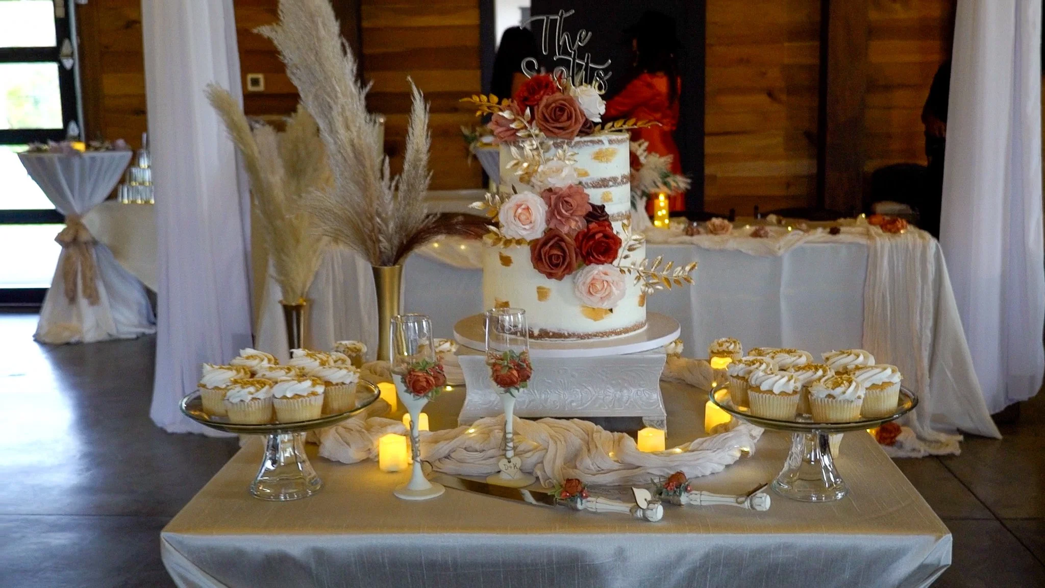 A wedding cake decorated with roses and gold accents, surrounded by cupcakes and candles on a table, with a rustic wooden interior in the background.