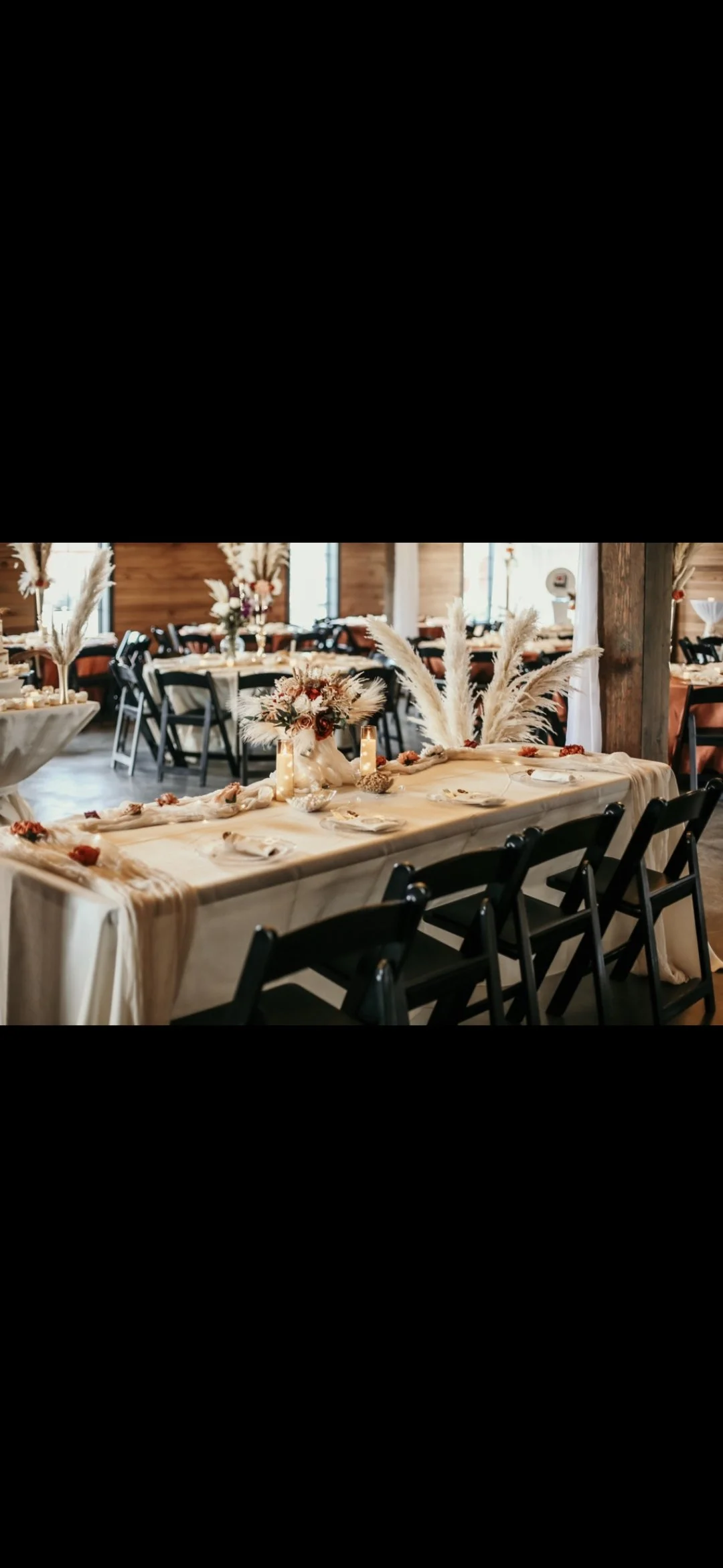 Bohemian wedding reception table decorated with pampas grass, dried flowers, candles, and neutral-colored tablecloth in a rustic venue with wooden walls and black chairs.