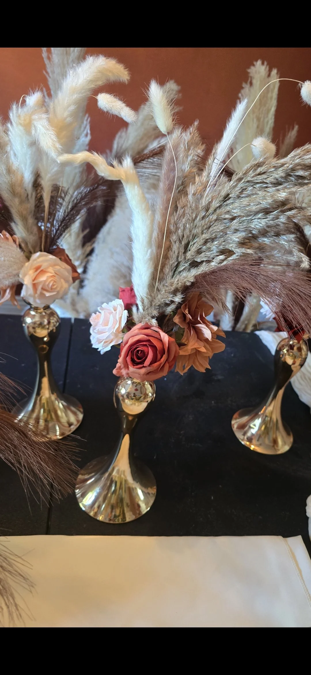 Decorative floral arrangement with pink and cream roses, beige and brown pampas grass, and other dried flowers in gold vases on a black table against an orange wall.